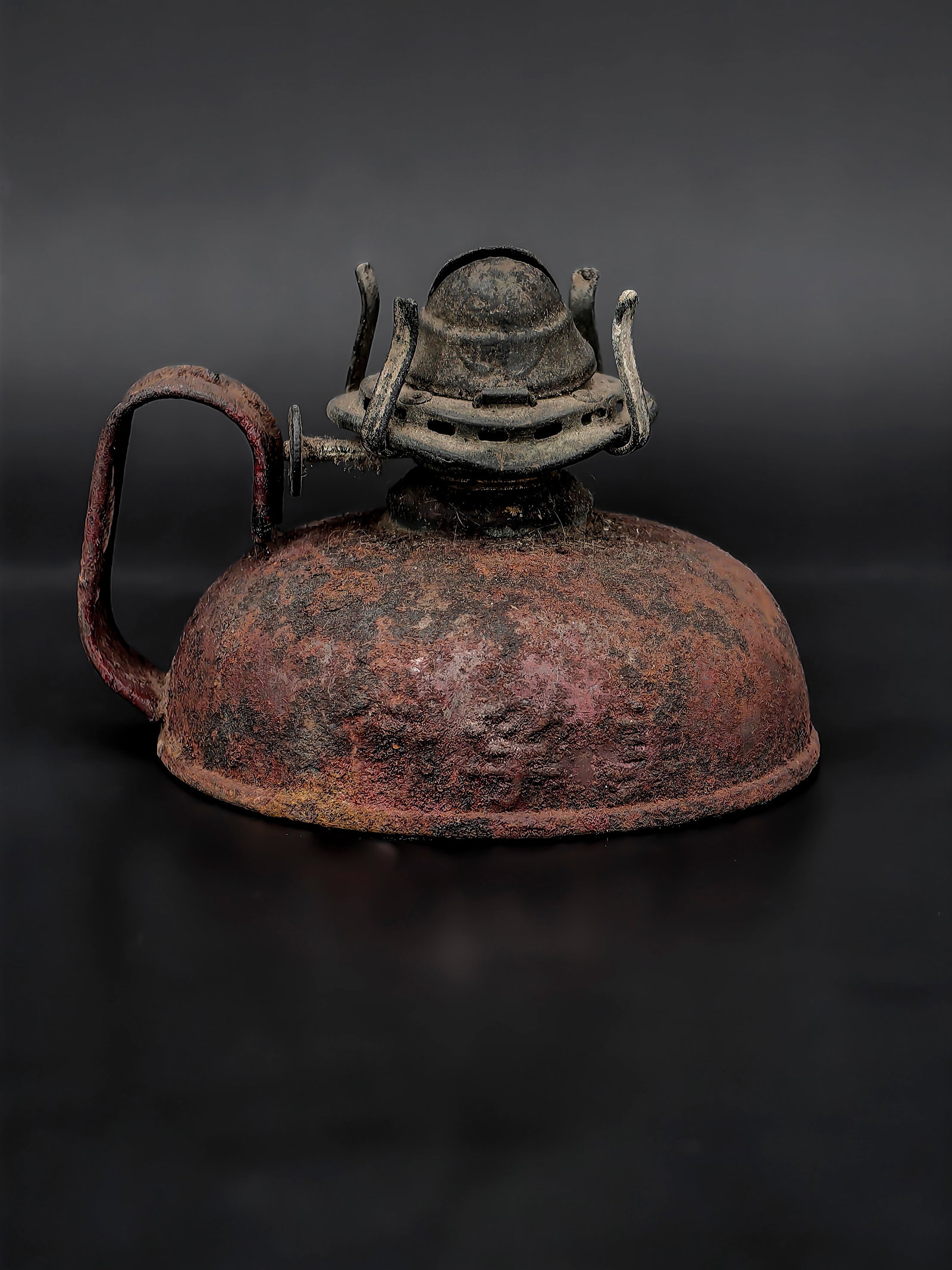 A Vintage Oil Lamp: A small vintage metal oil lamp featuring a rounded reservoir and fixed handle. The surface retains traces of original red paint beneath heavy oxidation, creating a richly textured patina consistent