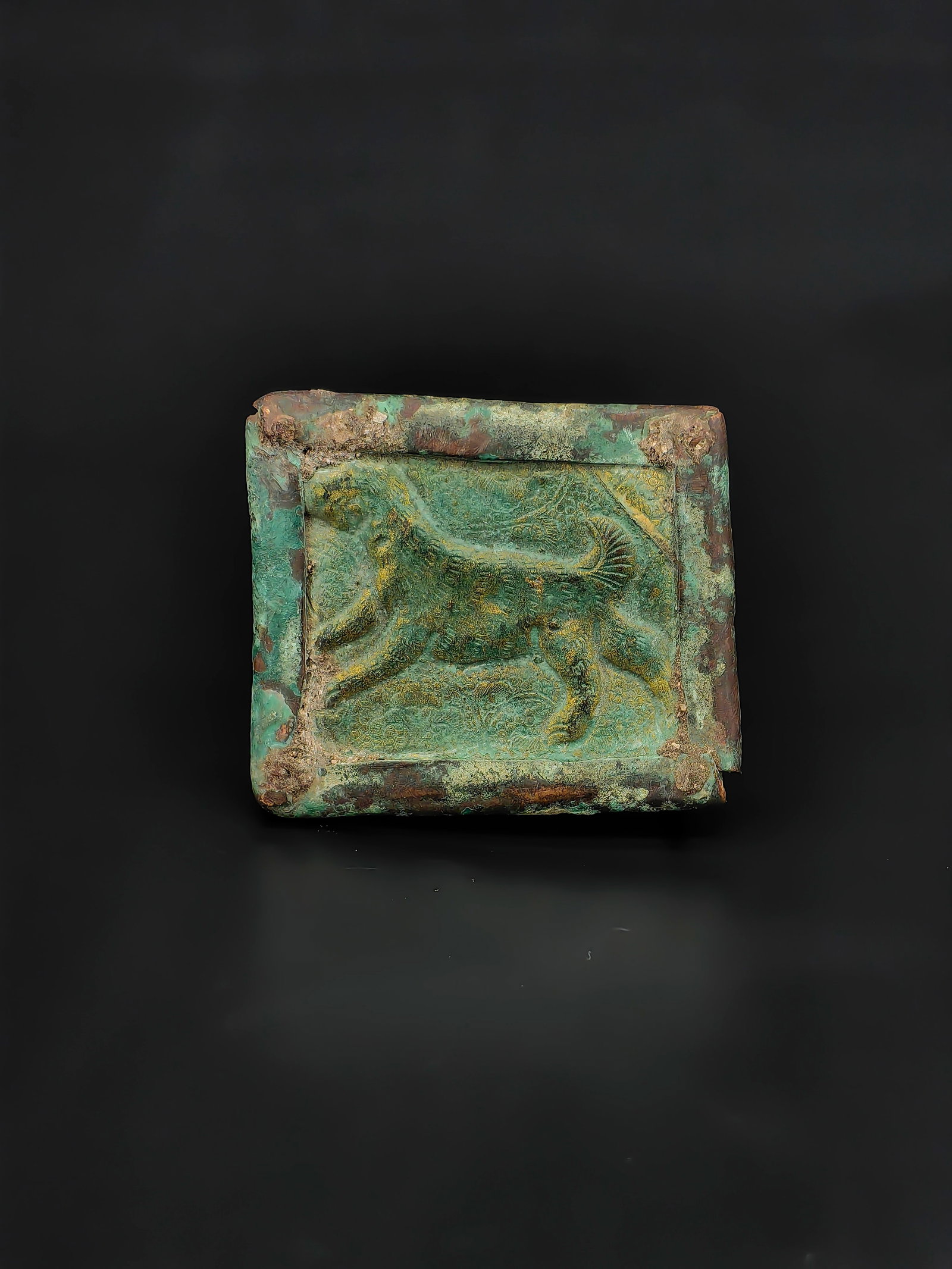 An Antique Chinese Bronze Plaque: A small rectangular bronze plaque featuring a raised relief of a running animal, possibly a deer or mythical beast, surrounded by a recessed border. The surface shows a natural green patina with