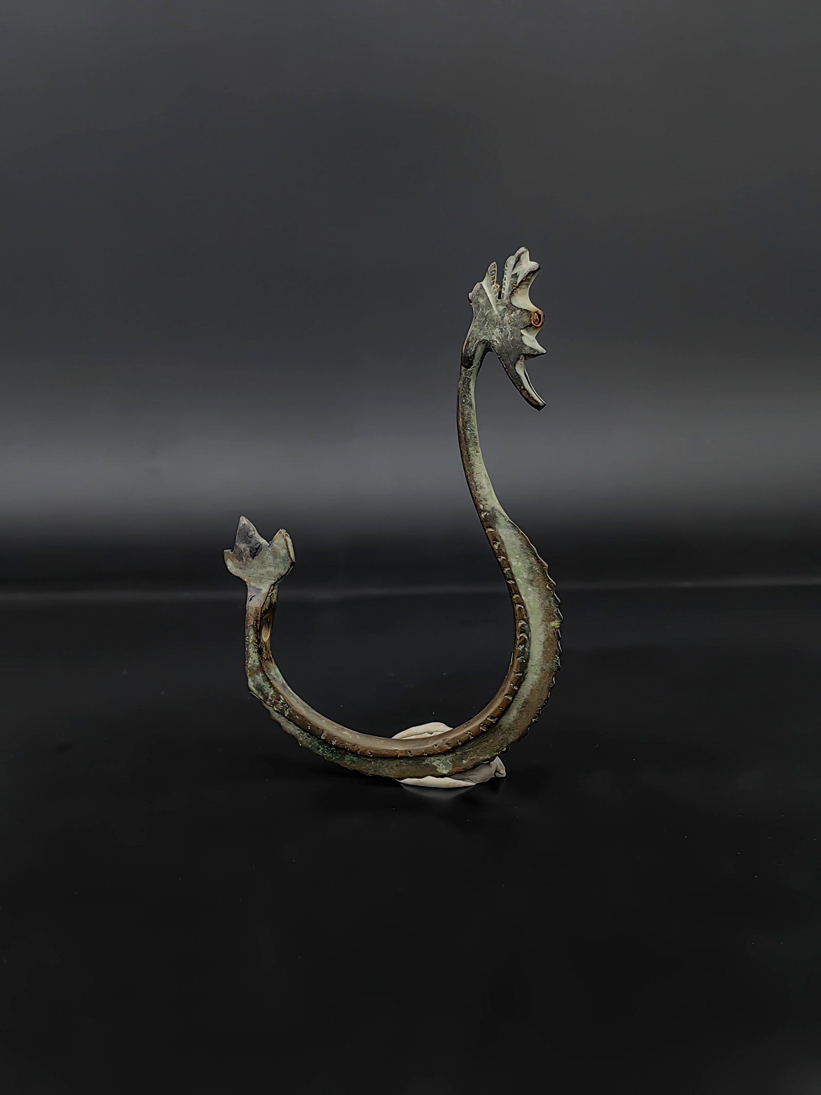 An Antique Chinese Bronze Belt Hook: A Warring States / Han Dynasty Bronze Dragon-Headed Belt Hook.this bronze belt hook features a gracefully curved, S-shaped body reminiscent of a swimming dragon. The head is meticulously cast into a p