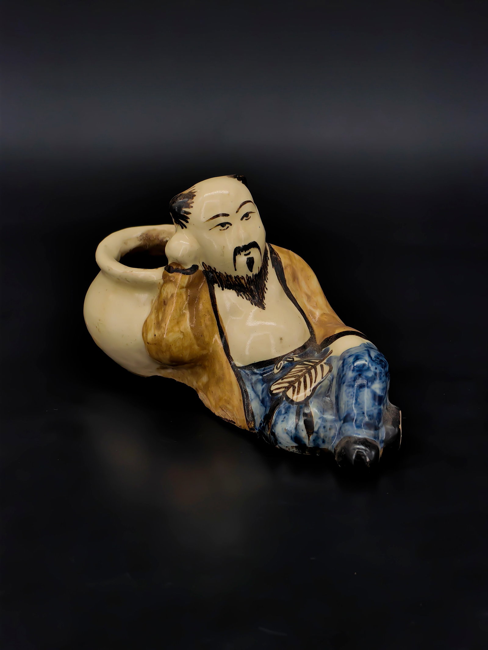 An Antique Shiwan Brown and Blue Enamel Figure-shaped Water Pot: This Republic period ceramic water pot is modeled as a reclining scholar with a lively facial expression, his beard and hair detailed in black pigment. He wears a brown-glazed wrap robe and trousers