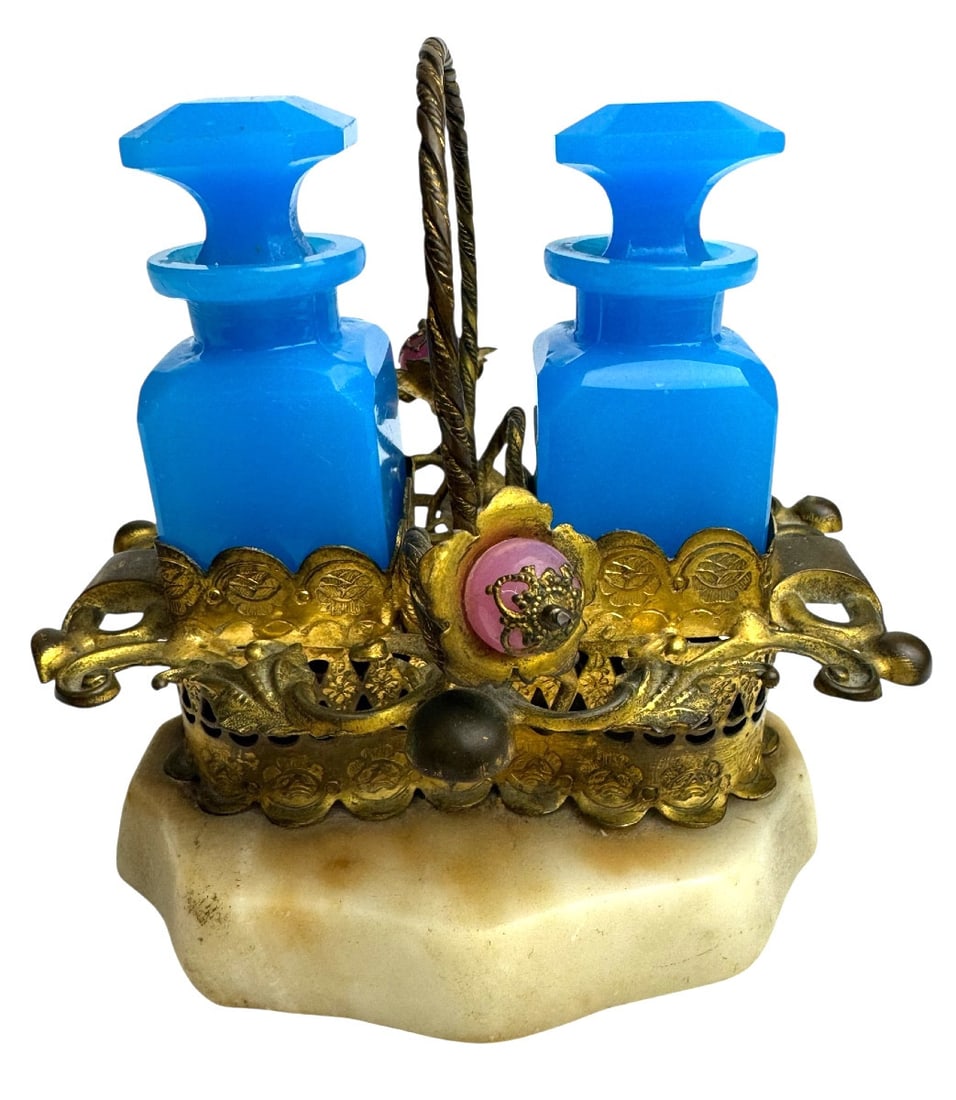 19th century Opaline perfume case with pomponne mount: A perfume case featuring two blue opaline glass bottles with their stoppers, mounted on a pompon-style base (scrolls adorned with leaves, flowers, and pink peppercorns) on a white marble surface. In v