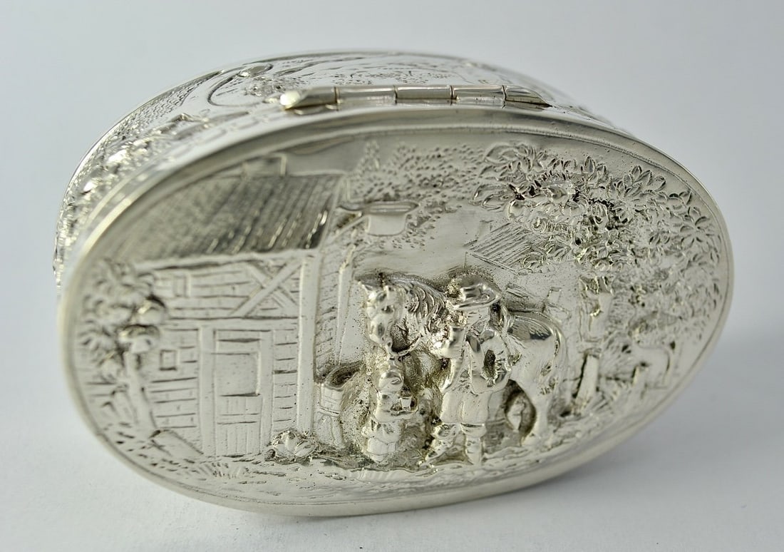 Oval Silver Box, Germany, Circa 1900: Oval-shaped box with a flat base, made of repoussé, chased, and guilloché silver.The hinged lid features figures in low relief: a standing man holding a tankard, a servant with a pot, and a horse ag