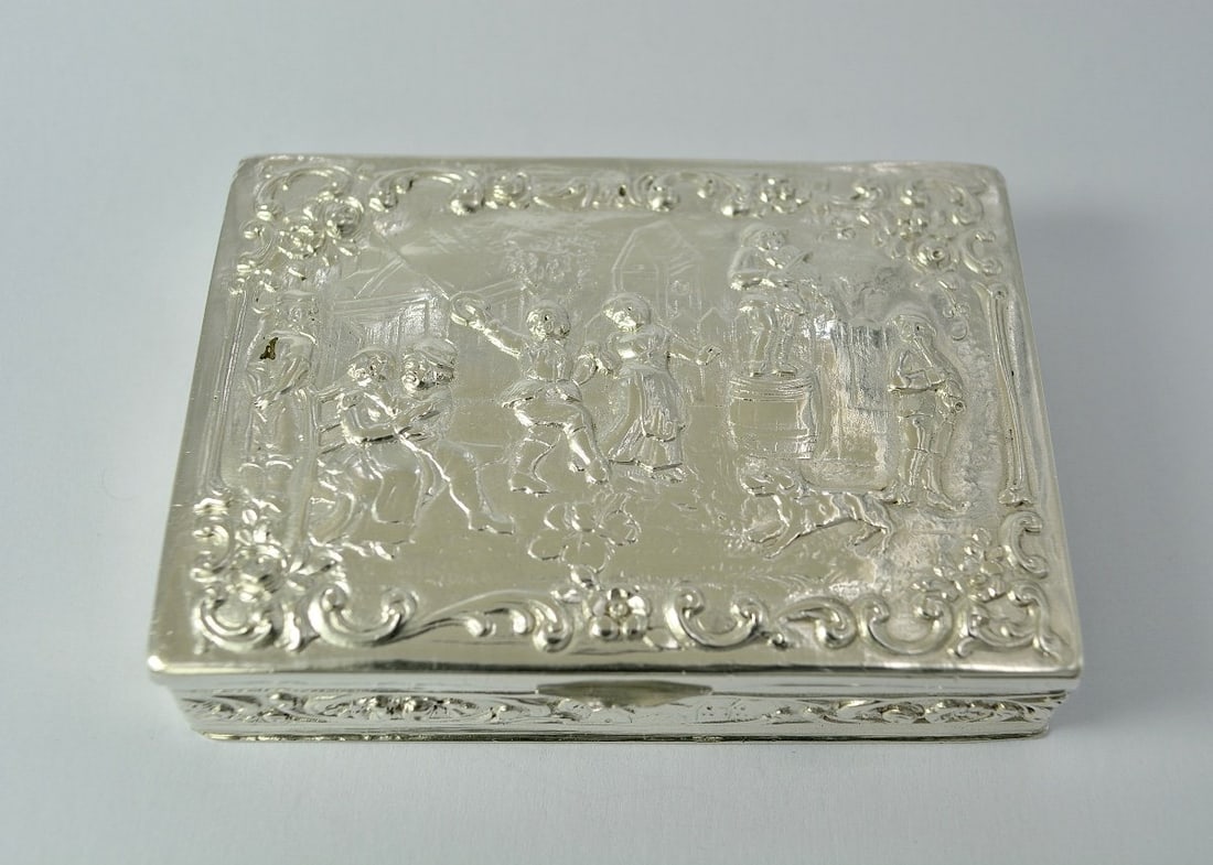 Silver Box, Germany, 19th Century: Rectangular repoussé silver box with a flat base. The hinged lid is decorated with a village festival scene within a frame embellished with rocaille motifs and flowers.Germany, 19th century.800/1000