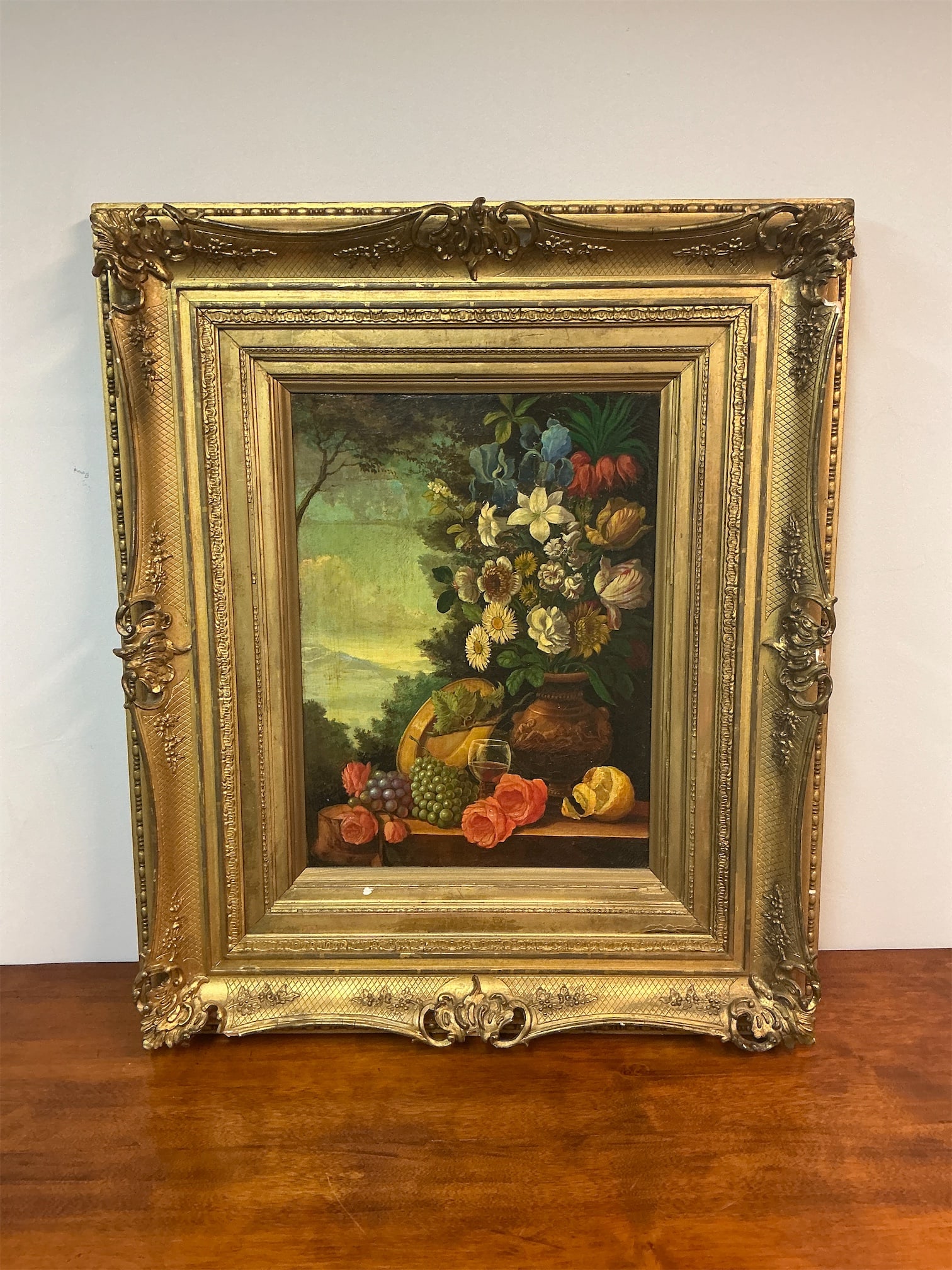 Antique framed oil on canvas still life: Antique framed oil on canvas still life features a vibrant arrangement of flowers and fruits, set against a serene landscape backdrop. The composition showcases an array of colorful blossoms,