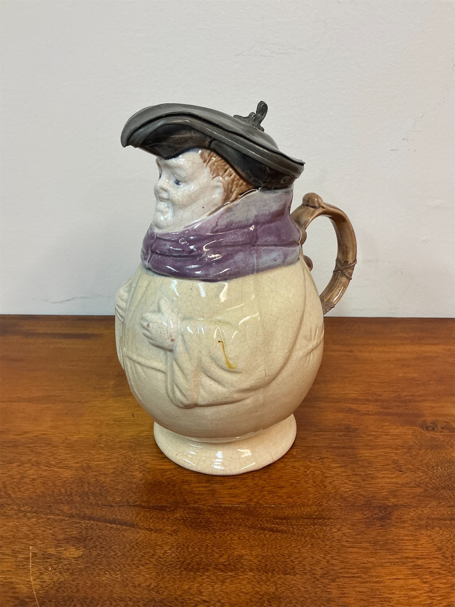Antique Majolica Monk Toby Pitcher: Majolica Monk Toby Pitcher showcases a charming design featuring a jovial monk with a striking purple scarf and a distinctive hat. Crafted with attention to detail, the pitcher features a sturdy