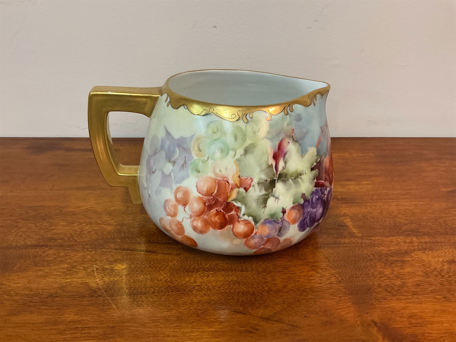 Antique Austrian Hand-painted Cider Pitcher: Austrian hand-painted cider pitcher features a charming depiction of lush grapes in vibrant colors. The elegant piece boasts a unique shape with a gracefully curved body and a stylish golden handle.