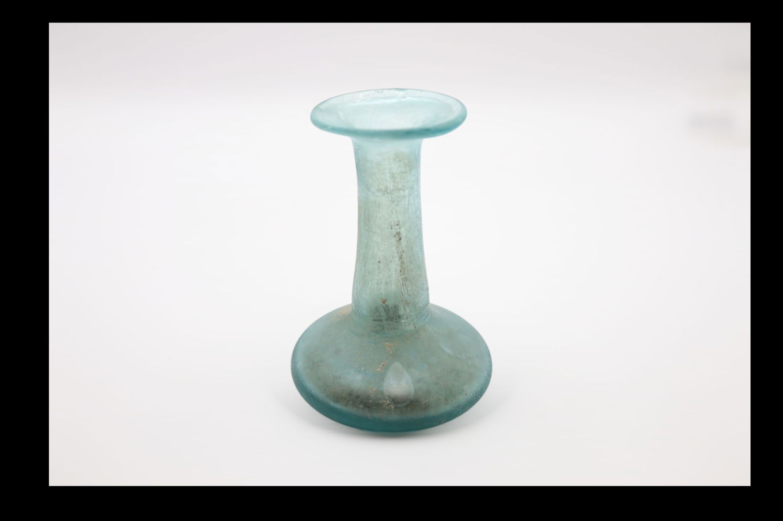 Roman Glass Perfume Bottle, Circa 1st Century AD: Translucent pale blue green. Rim folded out, over, and in, flattened on top with slight constriction to mouth; cylindrical neck with tooling indents around base; piriform body; thick, slightly concave