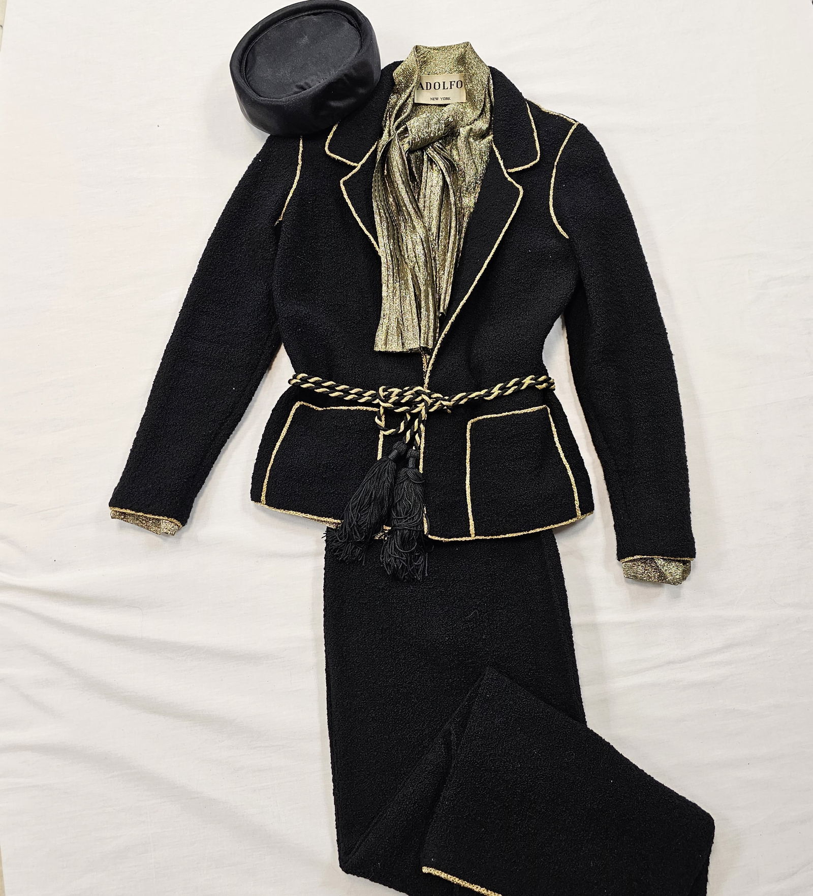 Adolfo Black Knit Pantsuit with Gold Lame Details: Vintage Adolfo black knit pantsuit with gold lame trim, a gold lame blouse with pleated tie, gold and black tasseled belt, and black silk pill box hat.Approximate measurements are: jacket - 24 in leng