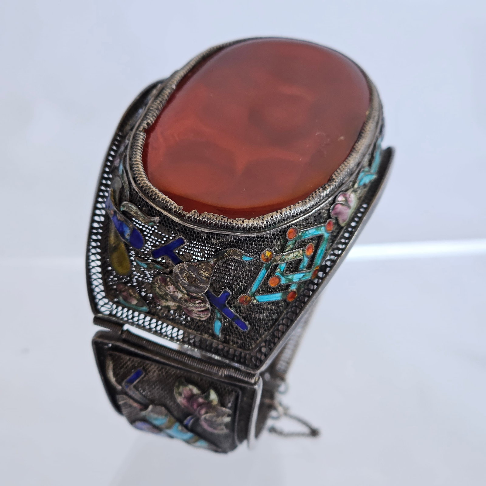 Chinese Export Carnelian, Enamel, Silver Bracelet: Centering an oval carnelian agate plaque measuring approximately 54.40 x 32.30 mm (2.14 x 1.27 in), highlighted by polychrome enamel, set in a silver filigree hinged bracelet mounting completed by a t