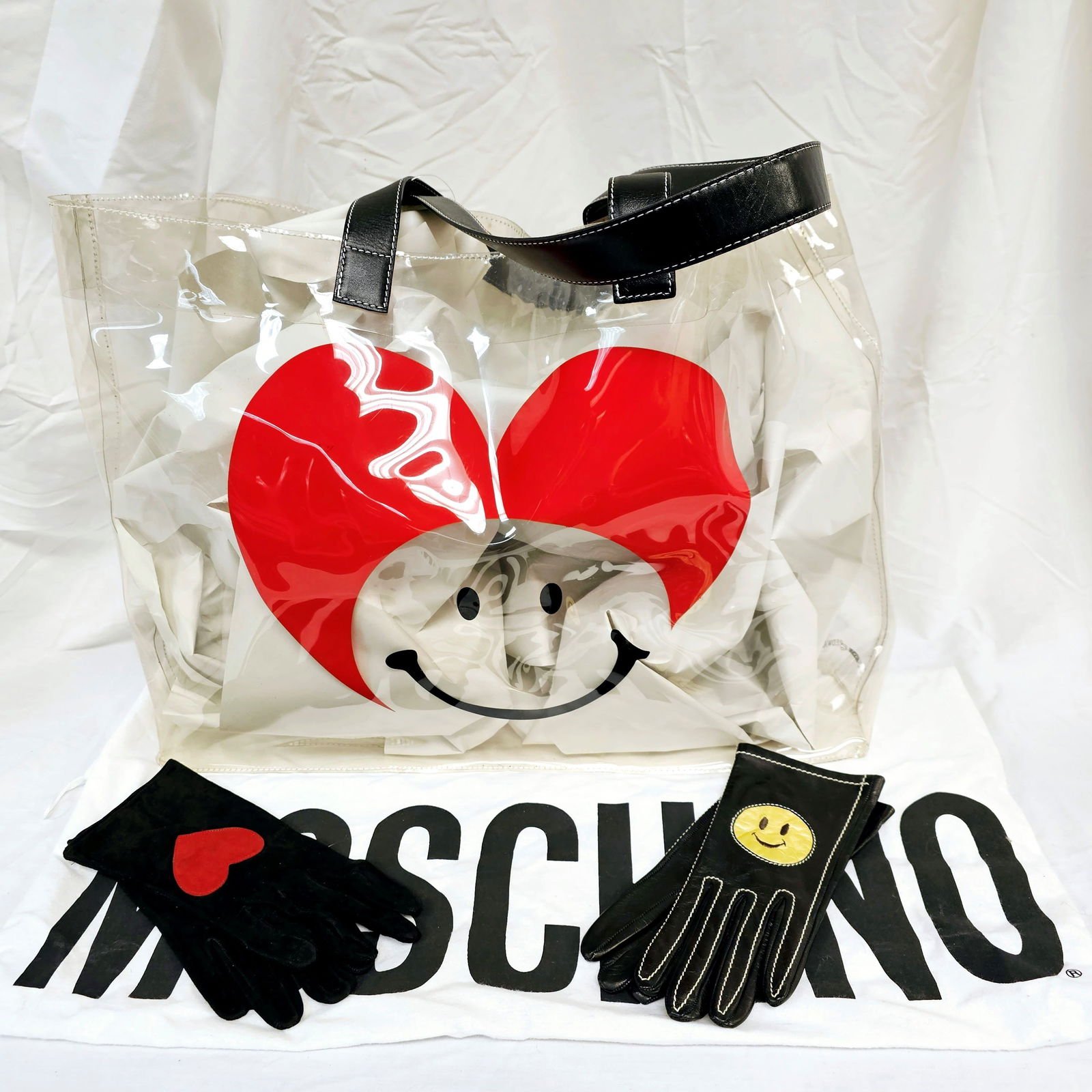 Grouping of Moschino Items: Grouping of Moschino items including a Moschino clear vinyl tote, a pair of black suede gloves with a red heart, and a pair of black leather gloves with white stitching and a yellow smiley face. 