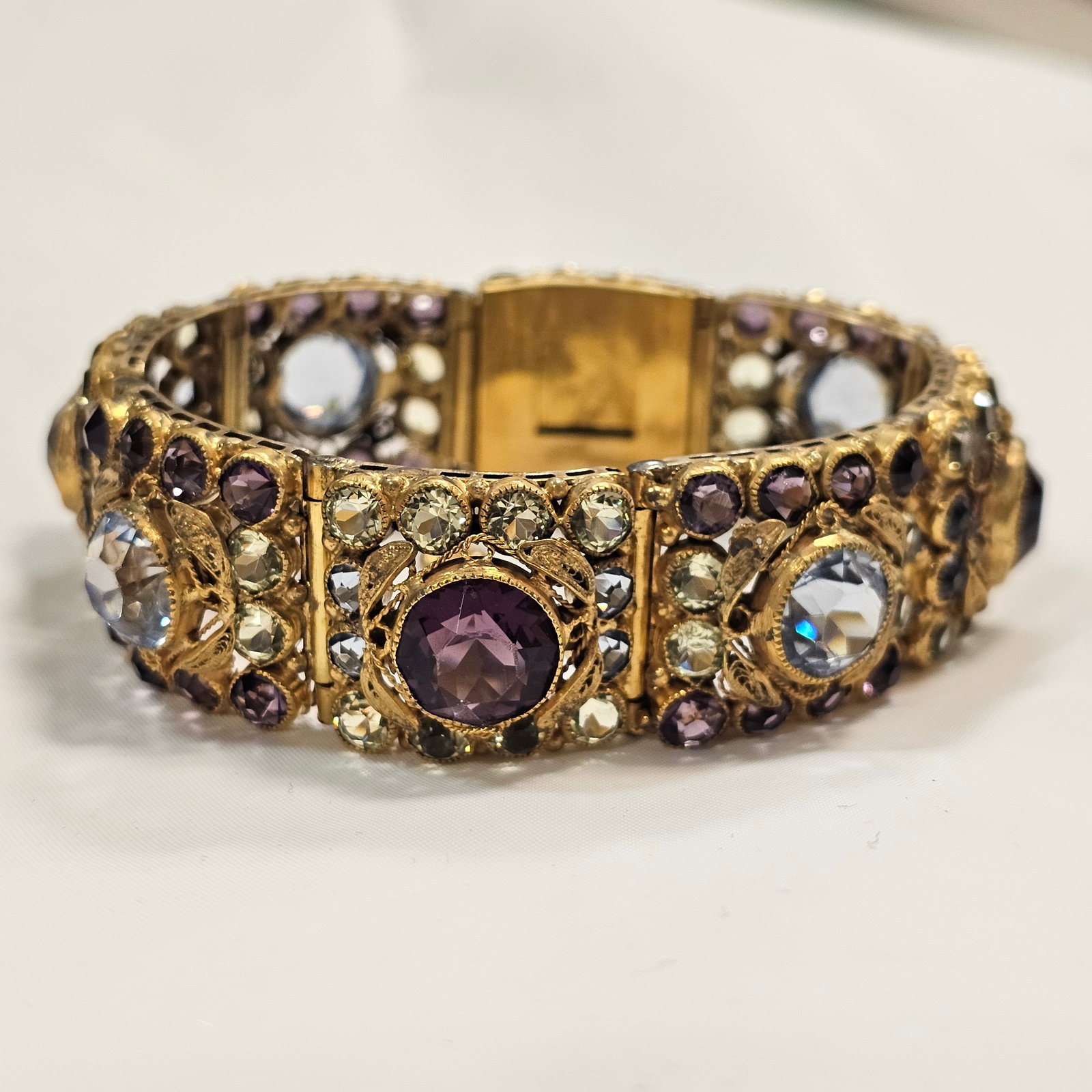 Hobe Rhinestone Articulated Bracelet: Goldtone filigree and alternating colors of purple and pastel blues and greens, larger faceted centers with smaller surrounding stones, each bezel set. Condition: some wear to goldtone,