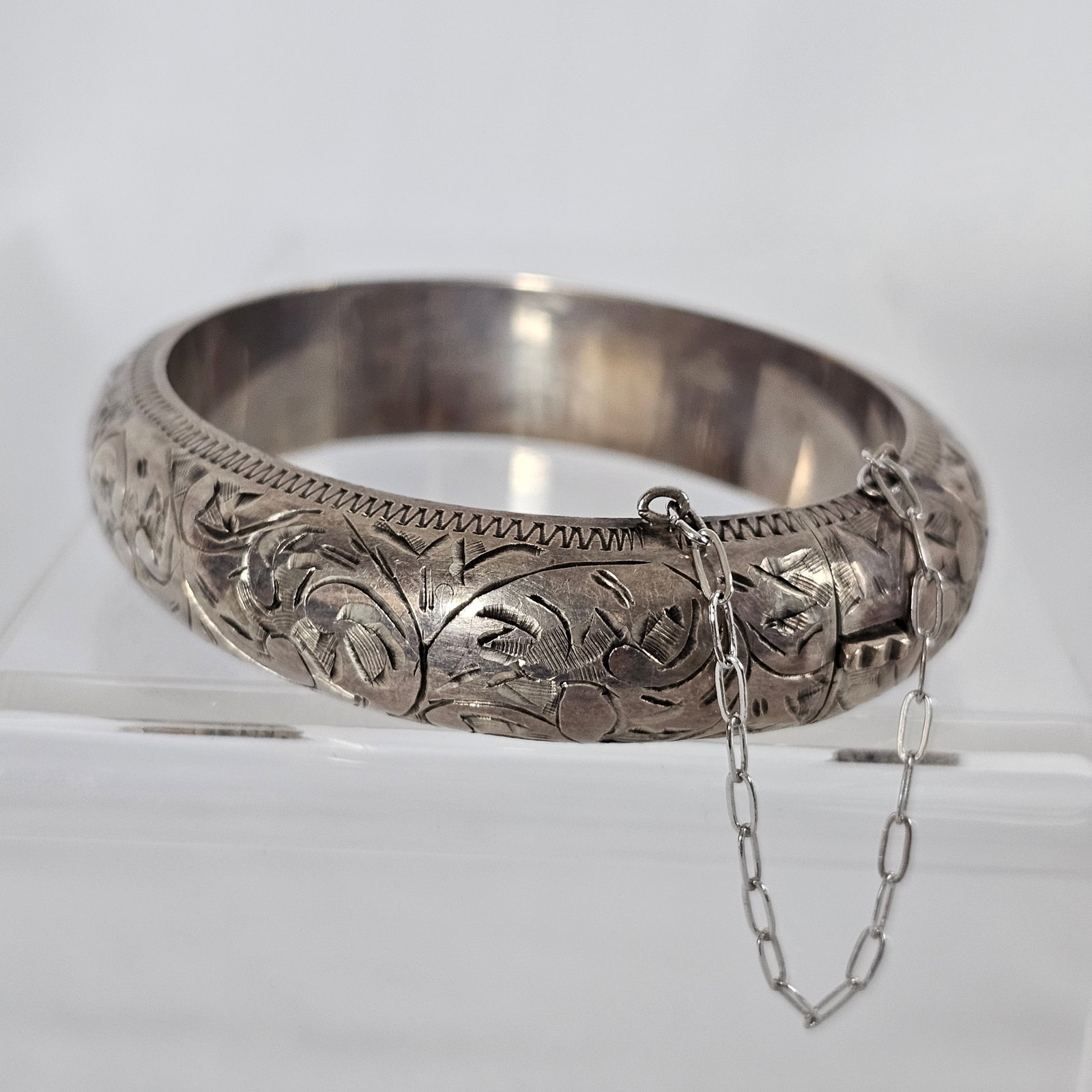 Sterling Silver Hinged Bangle Bracelet: The sterling silver hinged bangle bracelet measuring approximately 0.75 inches in width, completed by a tongue-and-groove clasp with safety chain, inner circumference 6 1/4 inches. Gross weight: 68.4 