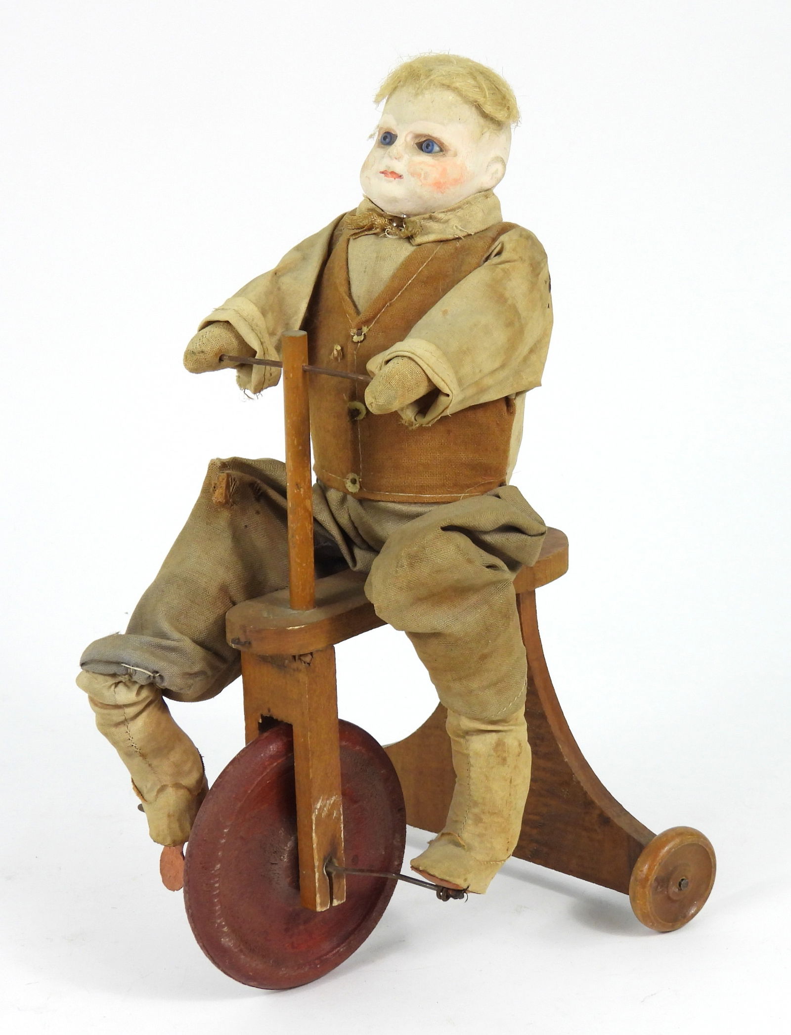 19C Bisque Head Bike Riding Boy Doll: 19th Century, Textile, wood, bisque. Depicts a blonde bisque head boy doll with blue glass eyes affixed to a wood tricycle with red wheels. Approx. 13 1/2" t x 7" w x 8" d