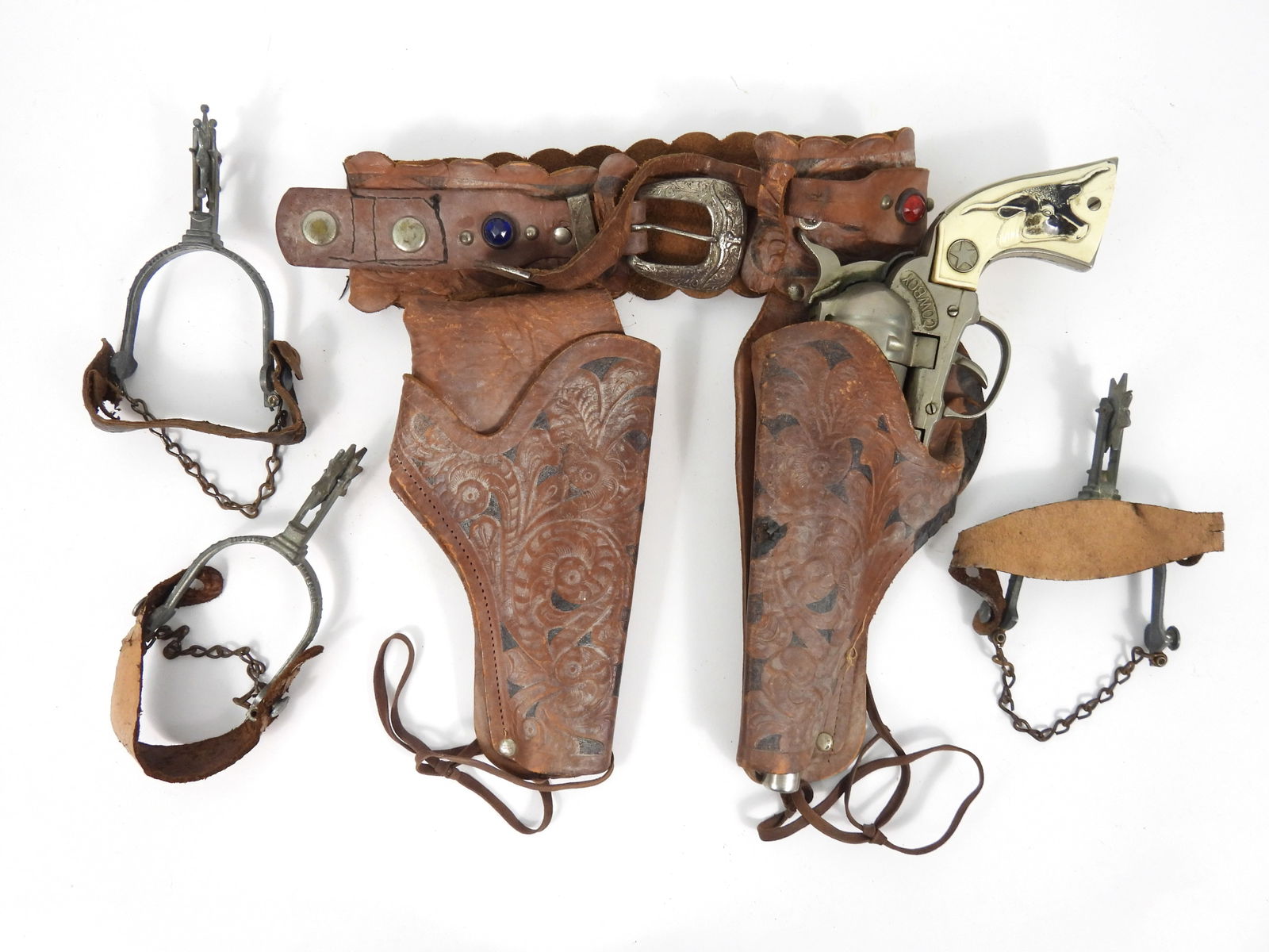 3PC Cowboy Cap Gun & Spurs: United States, 20th Century, Metal. Includes a Cowboy cap gun with black bull head handle, tooled leather and studded holster, and a pair of spurs. Cap gun 5" x 12 1/2", Spurs 5" x 2 1/2"