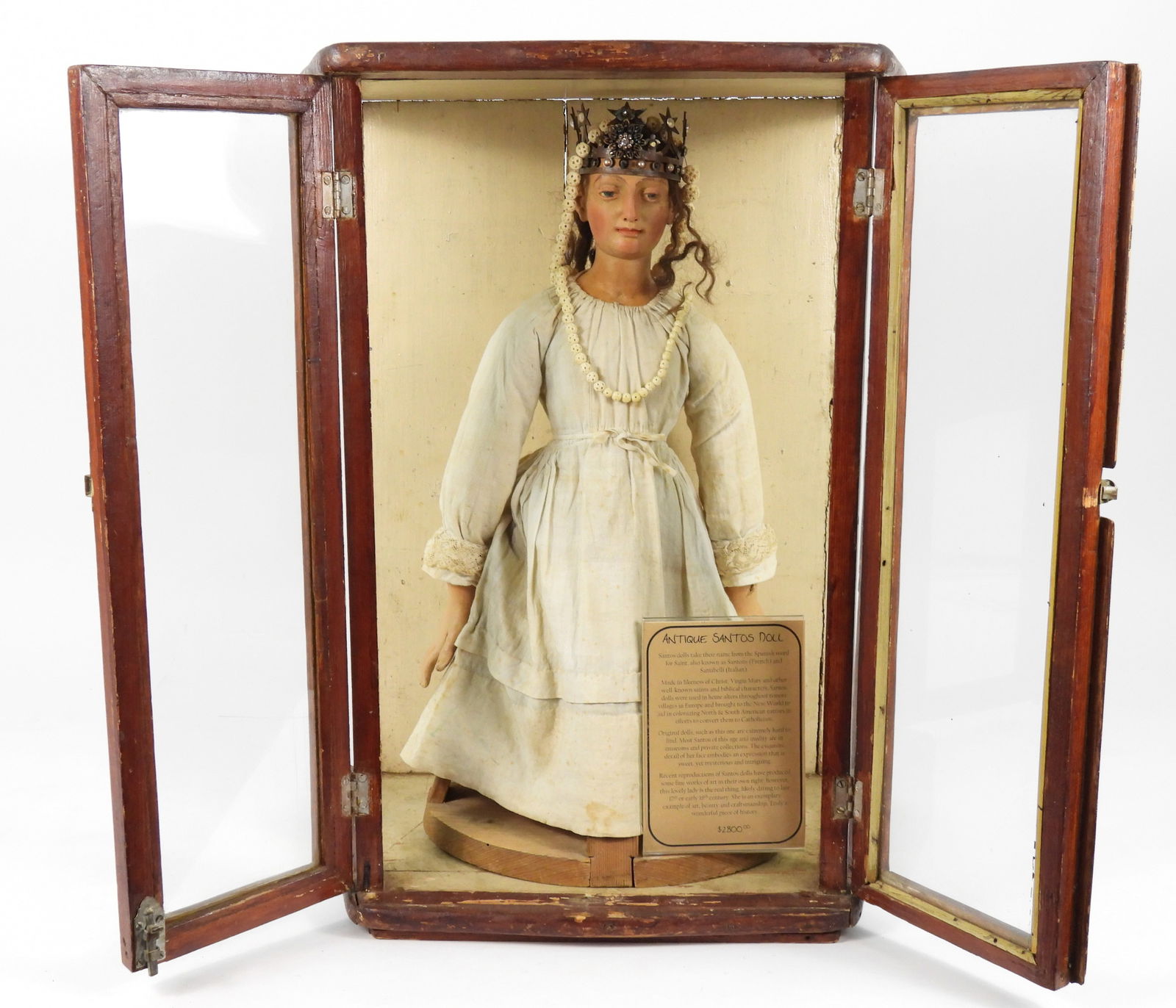 19C Santos Doll Display: South America, 19th Century, Wood, textile. Carved wood brunette Santos doll with painted blue eyes wearing a crown and layered white dress housed in a wood display case. Case 25 3/4" t x 20 1/4" w x
