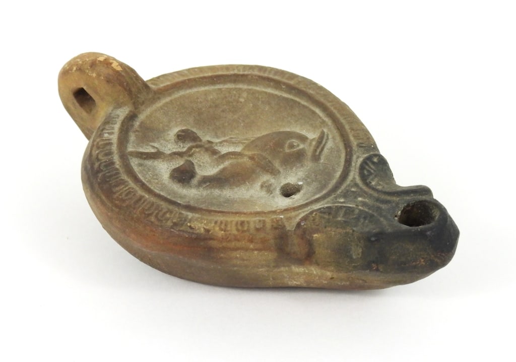 Ancient Roman Terracotta Oil Lamp: Cupid Eros riding a Dolphin in the discus. Spout is ornate with tapered nozzle wick hole. Appears to be 1st or 2nd century CE likely mold made. No maker marks observed. Measures 4" x 2.5" x 1.5" high.
