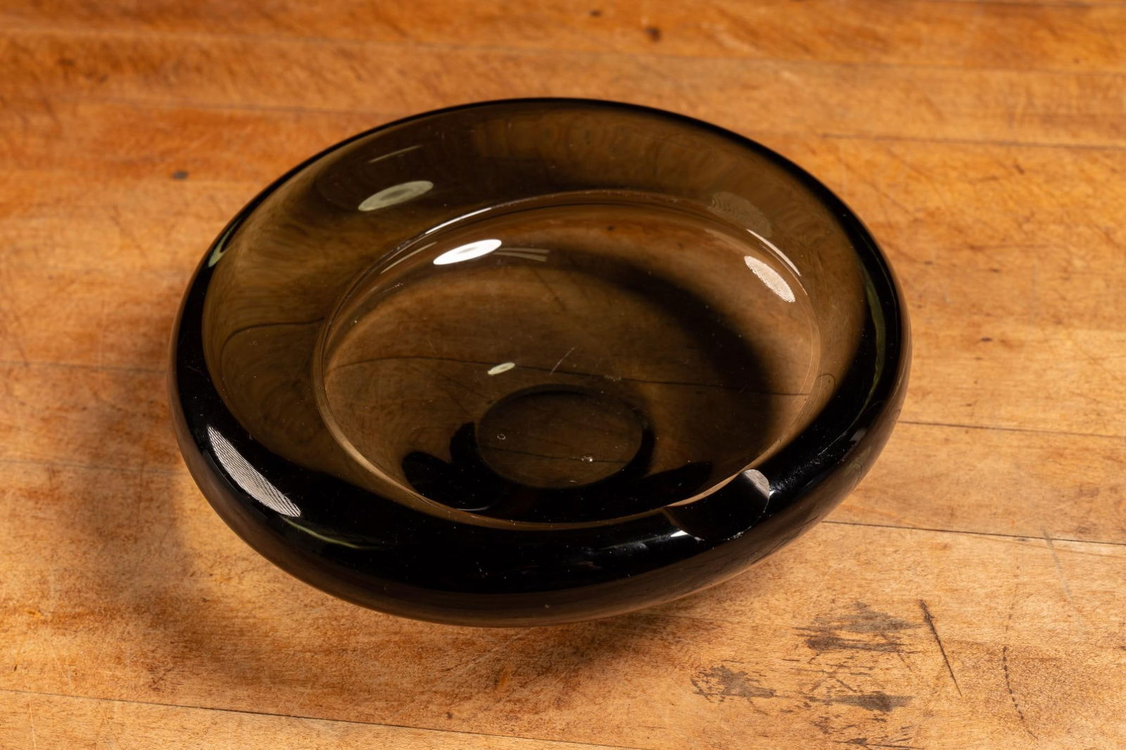 Smoked Glass Ashtray by Holmegaard: Darkened glass circular ashtray with a small ash receptacle. Appears to be free from cracks and chips, but exhibits minor scratches. 7" dia