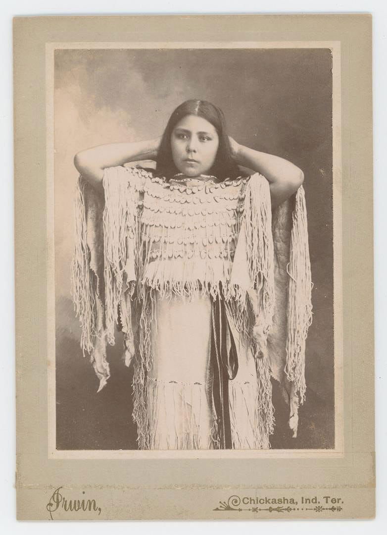 Kiowa Annie Indian Territory Cabinet Photo: Anna Berry posing with hands behind head and wearing fringed hide dress trimmed with elk teeth. "Anna E. Berry" variant personal names: Anna E. Jones Berry, Annie Berry, Dome-bay, Kiowa Annie. Taken b