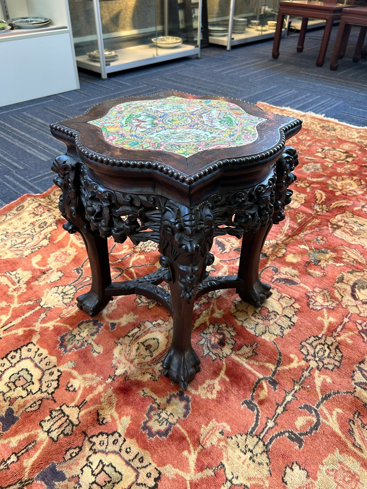 A Hongmu Stool with an Inset Famille Rose Plaque: The foliate porcelain top is decorated with flowers, birds, and antiques. The four legs are curved outwards and end in hoof feet, joined at the top with aprons. 17.3/4 in. high, 15 in. wide.