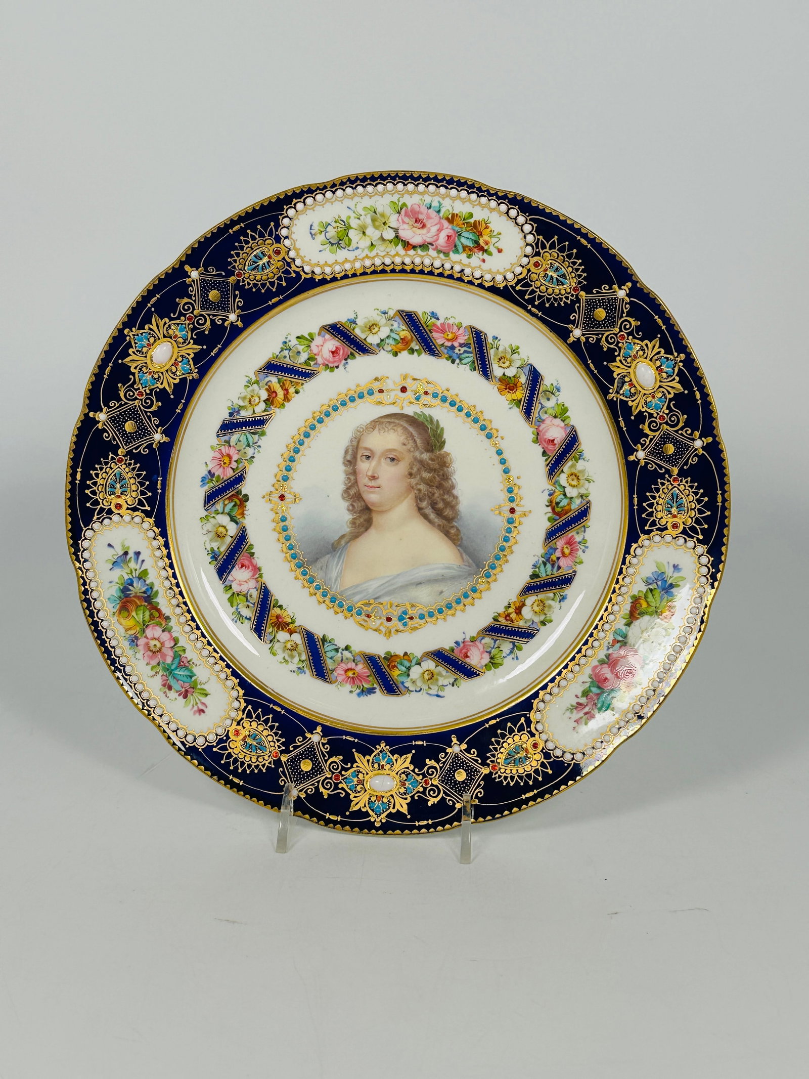 A Sevres Style 'Jeweled' Blue-Ground Portrait Plate: Spurious blue interlaced B's mark to the back, a shaped border featuring three oval reserves of flowers set on an intricately 'jewelled' band, depicting a bust-length portrait of a lady, framed by a '
