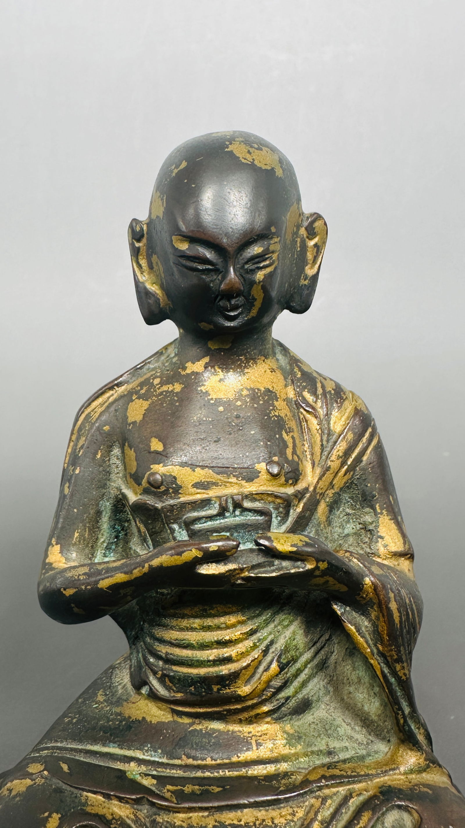 A Gilt-Bronze Figure of Seated Monk: Seated cross-legged, the robes cascade gracefully over the sides. The arms are held forward with hands gently crossed, the eyes lowered in serene meditation. 5.3/4 in. high, 4.1/4 in. wide.