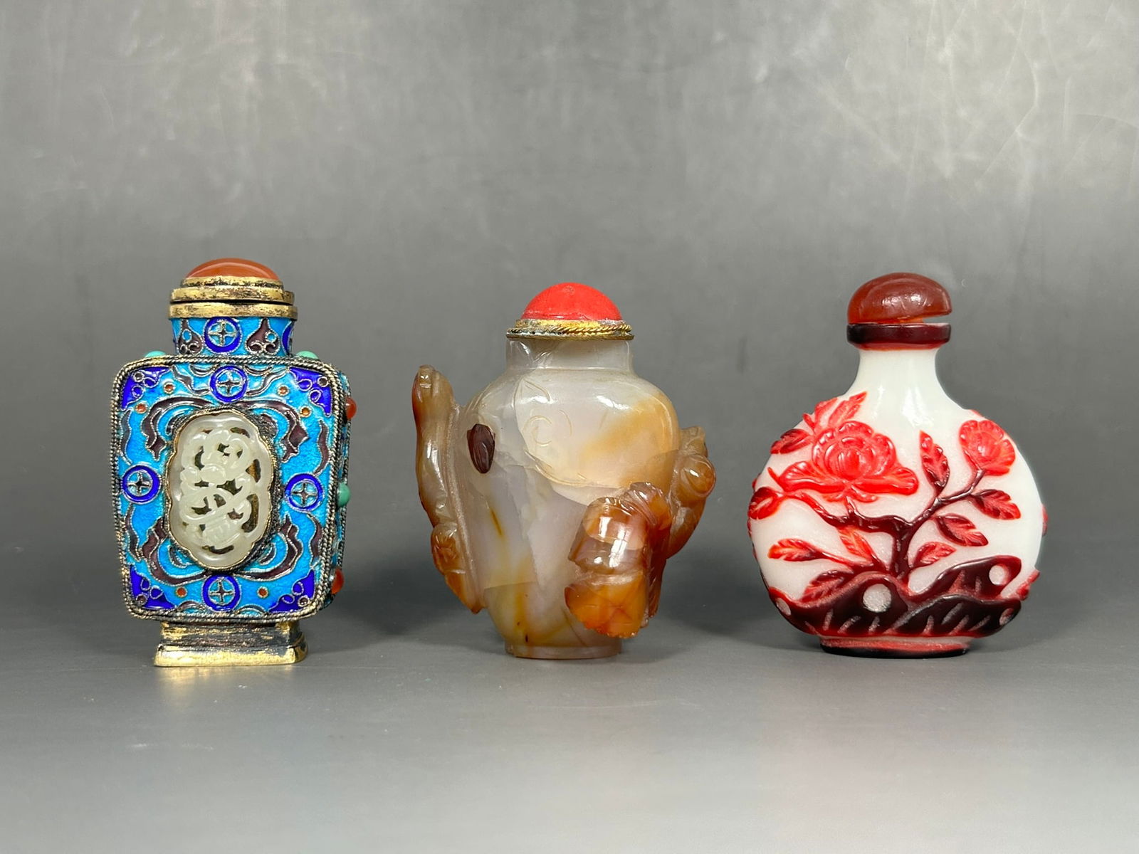 A Group of Three Chinese Snuff Bottles: Comprising a jade-inset cloisonne enamel snuff bottle, 2.3/4 in. high, 1.5/8 in. wide; a carved agate snuff bottle, 2.1/2 in. high, 2 in. wide; and a red-overlay glass snuff bottle, 2.1/2 in. high, 2