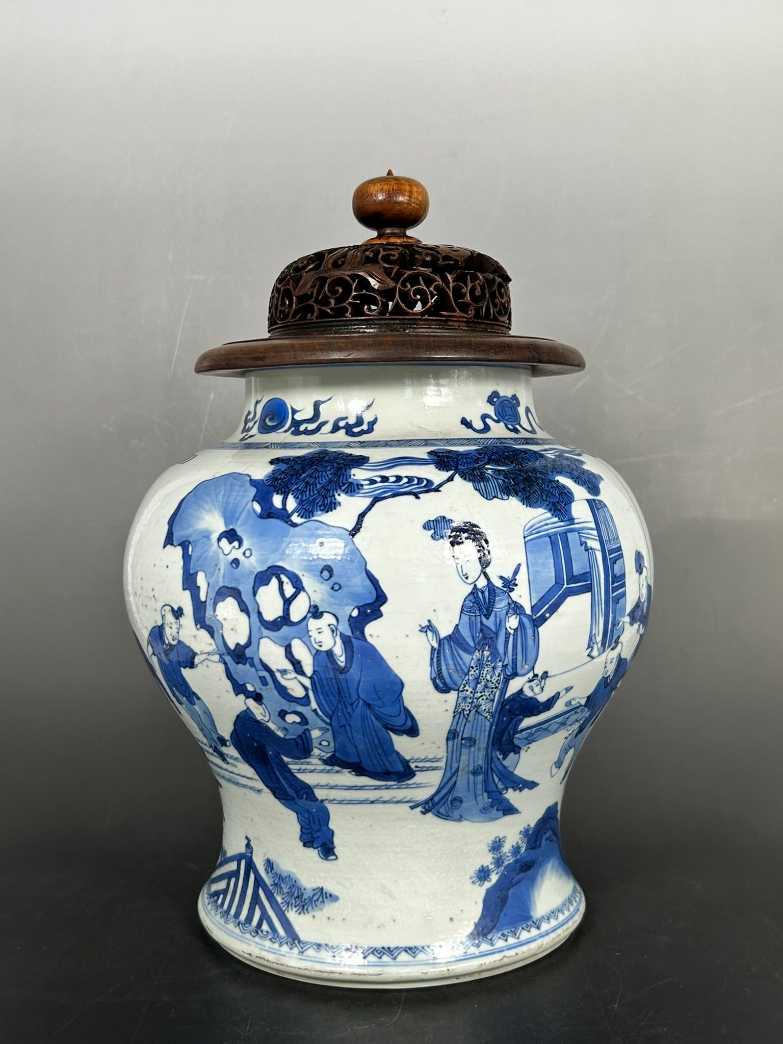 A Chinese Blue and White 'Ladies and Children' Jar and Wood Cover, Kangxi Period, Qing Dynasty: 12.3/4 in. high, 8.1/2 wide overall.