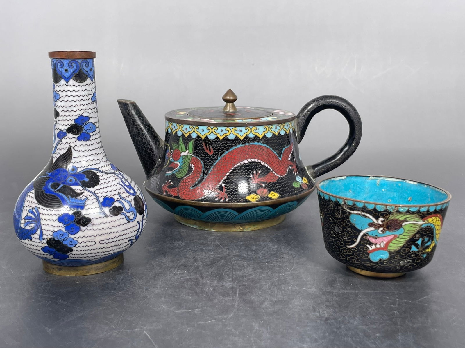 A Group of Three Chinese Cloisonne Enamel 'Dragon' Ware: Comprising a teapot and cover, 3.7/8 in. high, 7 in. wide; a cup, 1.7/8 in. high, 2.7/8 in. wide; and a small vase, 5 in. high, 3 in. wide.