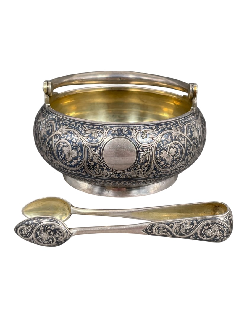 Doyle Provenance: A Russian Parcel-Gilt Silver and Niello Sugar Bowl and Sugar Tong: A squat, circular vessel fitted with a swing handle and raised on a circular foot, decorated in niello with scrolling foliate motifs interspersed with vacant circular reserves. The tongs are of typica
