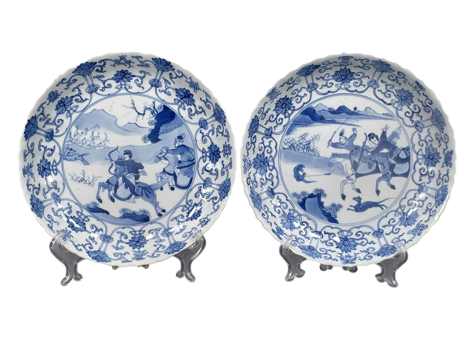 A Pair of Chinese Blue and White 'Hunting Scene' Porcelain Dishes, Kangxi Period, Qing Dynasty: Each depicting an equestrian couple beneath the Eight Buddhist Emblems (bajixiang), the reverse bearing an encircled six-character apocryphal Chenghua mark. 7.3/4 in. diam. each. A larger comparable l