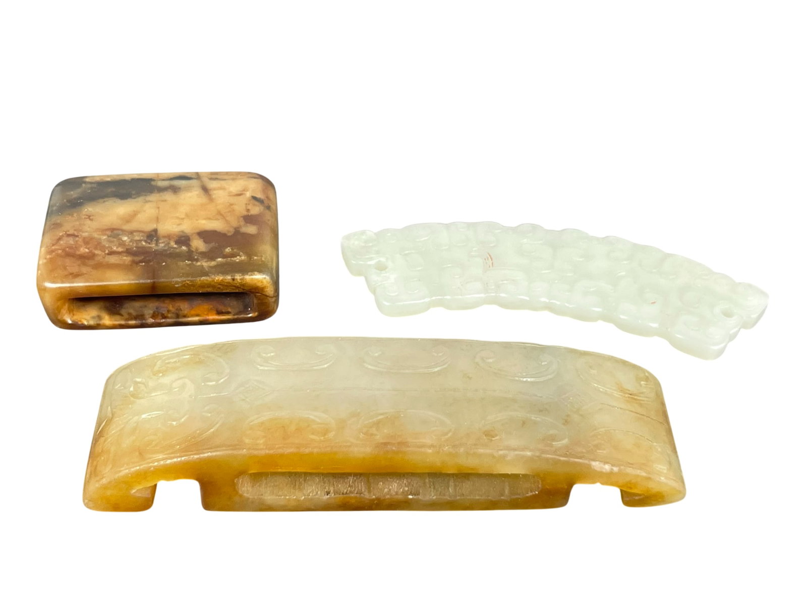 A Group of Three Chinese Carved Jade Objects: Comprising a belt buckle, 3.3/8 in. long, 7/8 in. wide; a pendant, 2.7/8 in. long, 3/4 in. wide; and a small belt buckle, 1.1/2 in. long, 1 in. wide.