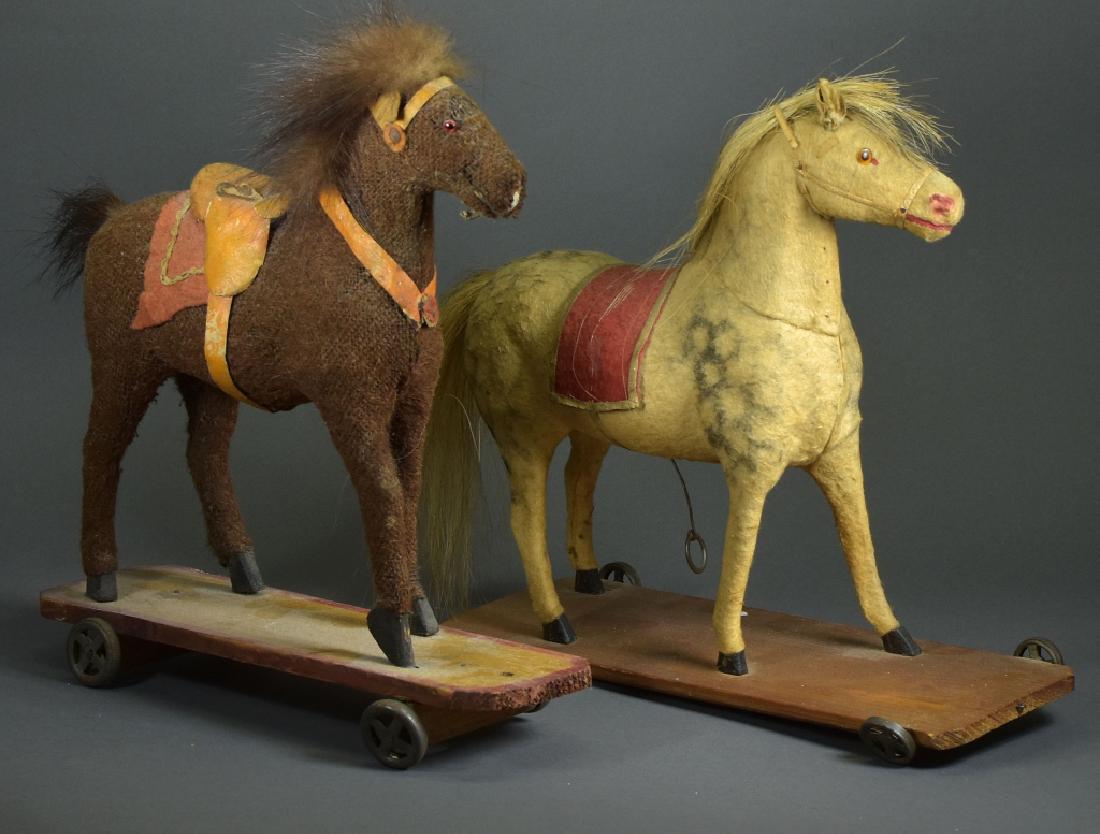 Two Pull Toys: Two horses, toys on wheeled bases. Brown is burlap covered with glass eyes, fur mane and felt saddle. Second is felt covered with horse hair mane, both unmarked, tallest is 11". Metal wheels.