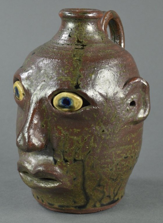 Anita Meaders Georgia Face Jug: Slightly open mouth with no teeth. Having olive green drip glaze over brick red glaze. Blue eyes. Thin delicate handle. Signed on base, “Meaders Pottery Anita Meaders.” 7 1/2” H.