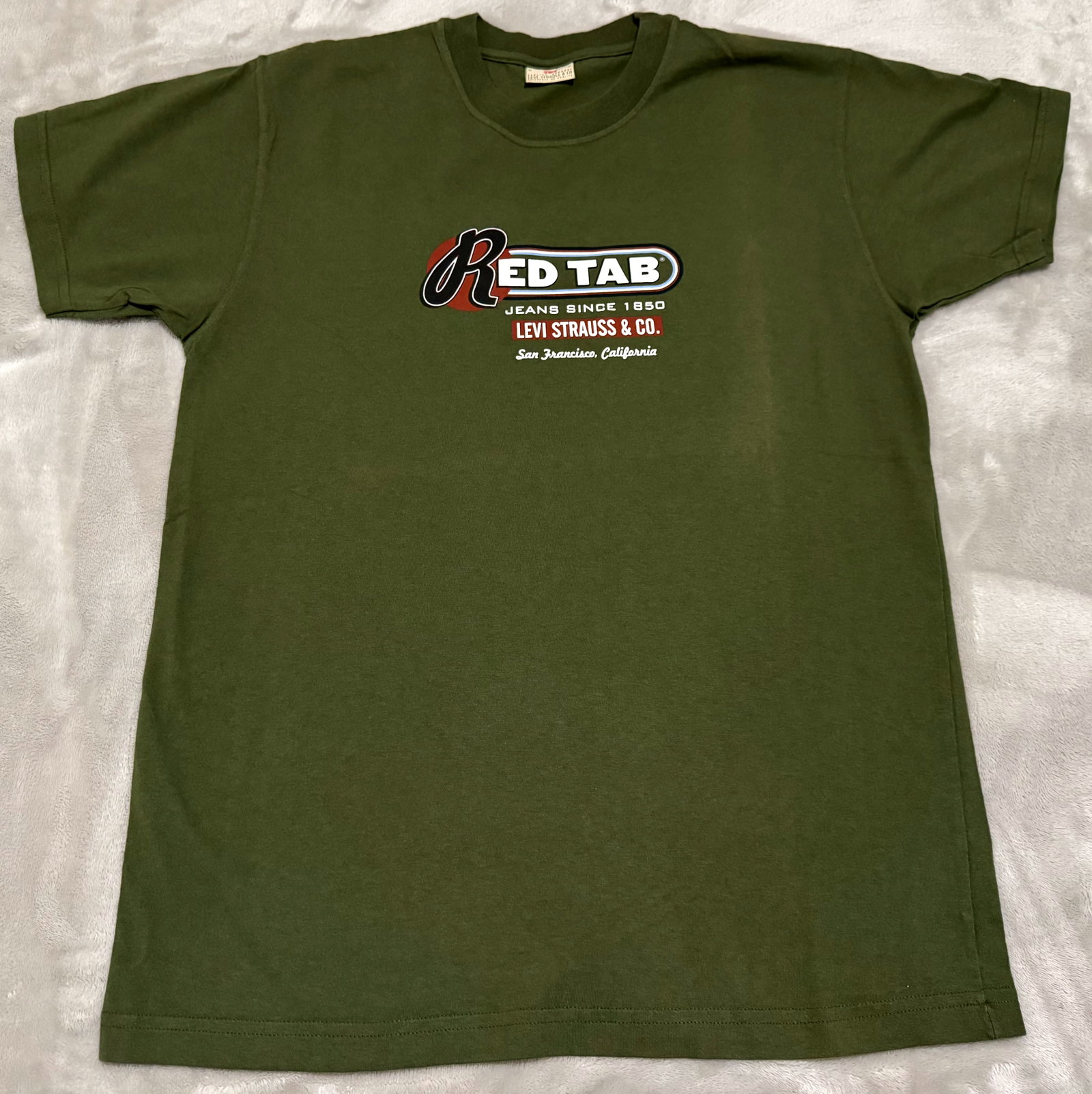 NOS 90s Vintage Levis Olive Green Red Tab Jeans Graphic Tee Shirt: Unused Features a sharp multi-color chest print highlighting the iconic "Red Tab" line with classic "Jeans Since 1850 / Levi Strauss & Co. / San Francisco, California" retro script typography. True vi
