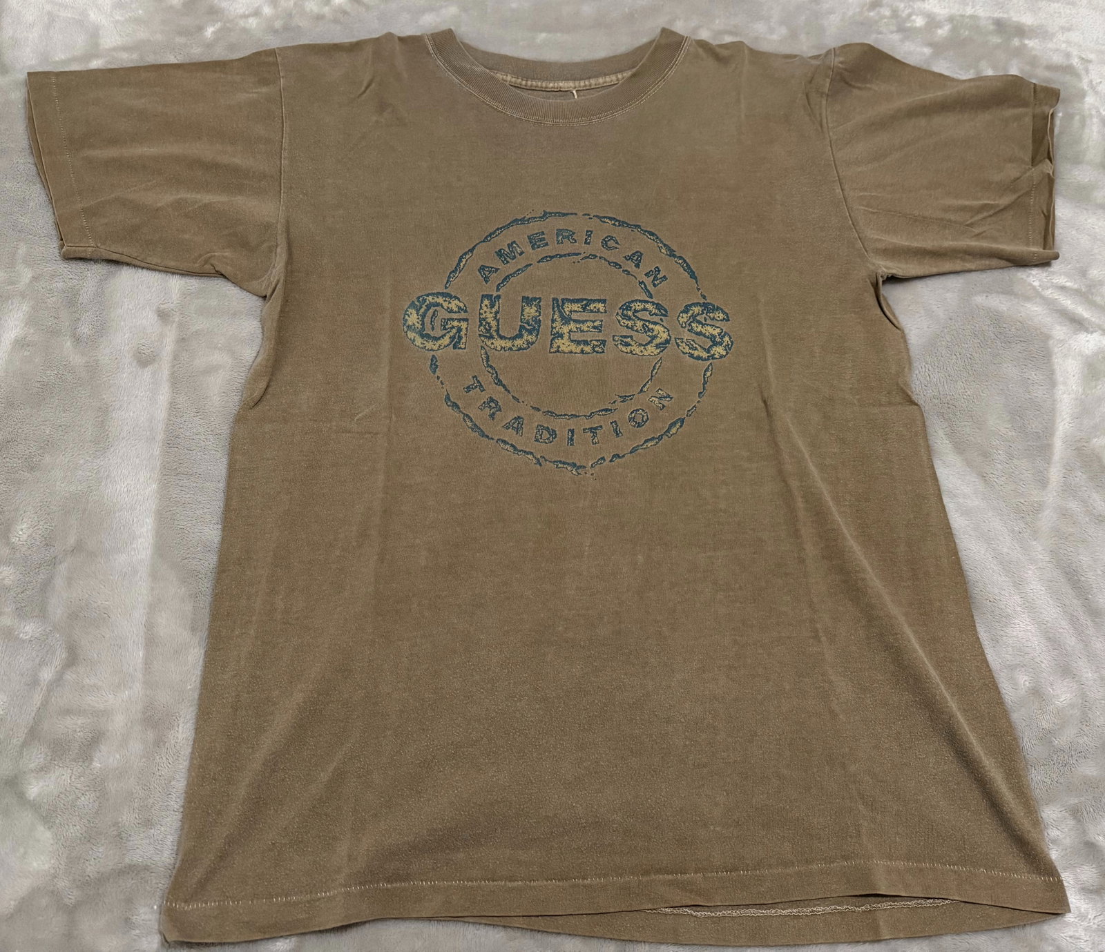 90s Vintage Guess American Tradition Khaki Tan Tee Shirt: Highly collectible dual-sided retro piece. The front features a large, heavily distressed circular chest graphic reading "AMERICAN GUESS TRADITION" in a textured blue and yellow fill. The upper revers