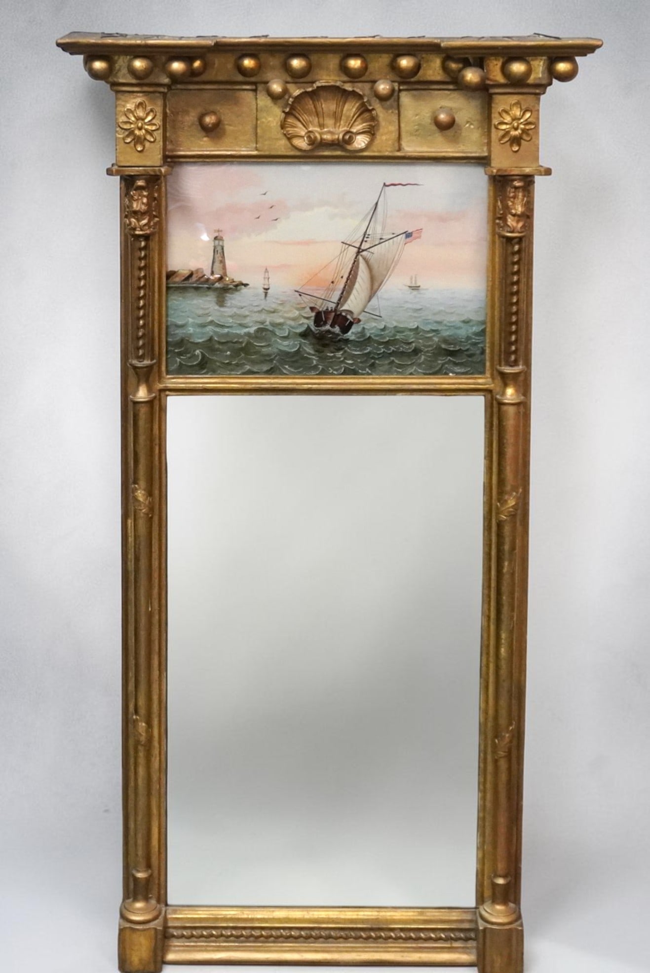 ANTIQUE AMERICAN 19th c GILTWOOD EGLOMISE MARITIME MIRROR: Antique 19th c American Federal lemon gilded mirror with upper eglomise panel depicting a sailing ship and lighthouse. With acorn finials and a central shell motif. Measures 20"x38"x5" Provenance: A W