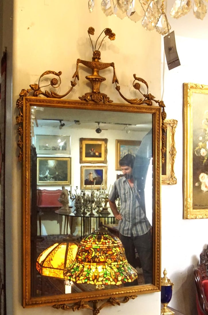 French Louis XVI Style Giltwood Urn Crest Mirror: An elegant French Louis XVI style giltwood wall mirror measuring 48 inches high by 26 inches wide. The rectangular frame features a classic beaded and leaf-tipped interior molding housing a large mirr