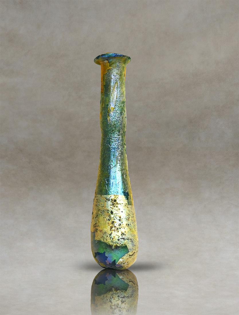 Roman Glass Unguentarium Perfume Bottle: Roman unguentary of pale colourless blown glass, probably used as a perfume bottle. The vessel has a flattened rim and a long, narrow cylindrical neck that widens towards the body. The lower part of t