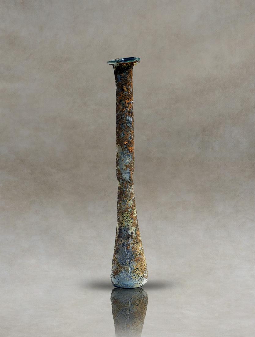Roman Glass Unguentarium Perfume Bottle: Roman unguentary of blue pale colourless blown glass, probably used as a perfume bottle. The vessel has a flattened rim and a long, narrow cylindrical neck that widens towards the body. The lower part