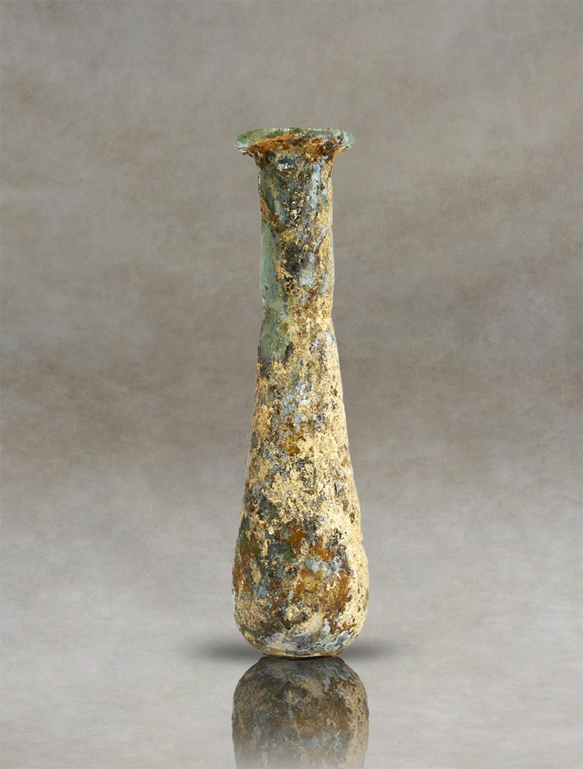 Ancient Roman Glass Unguentarium: An ancient Roman translucent-glass unguentarium. It has a pyriform body, with a cylindrical neck tapering at the base, and a bent and everted rim. The base is flattened. The vessel is filled with encr