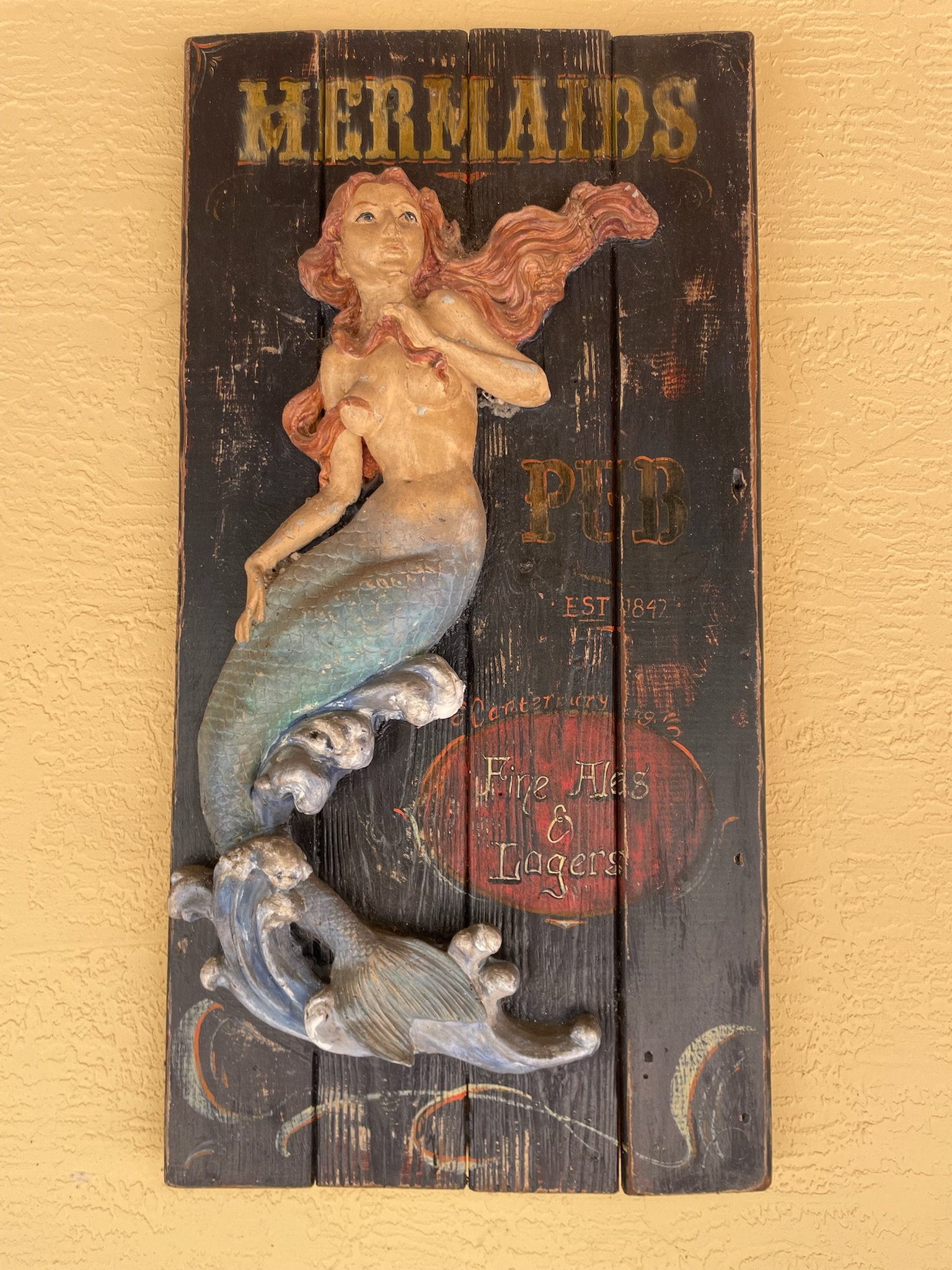 Mermaids Pub Sign: Fine Ales and Lagers. Wood with quality cement or plaster mermaid, handpainted. Dimensions: H 43" x W 22" x D 6" Minor signs of hanging in a covered porch. Low Estimate: 300.00 High Estimate: 500.00