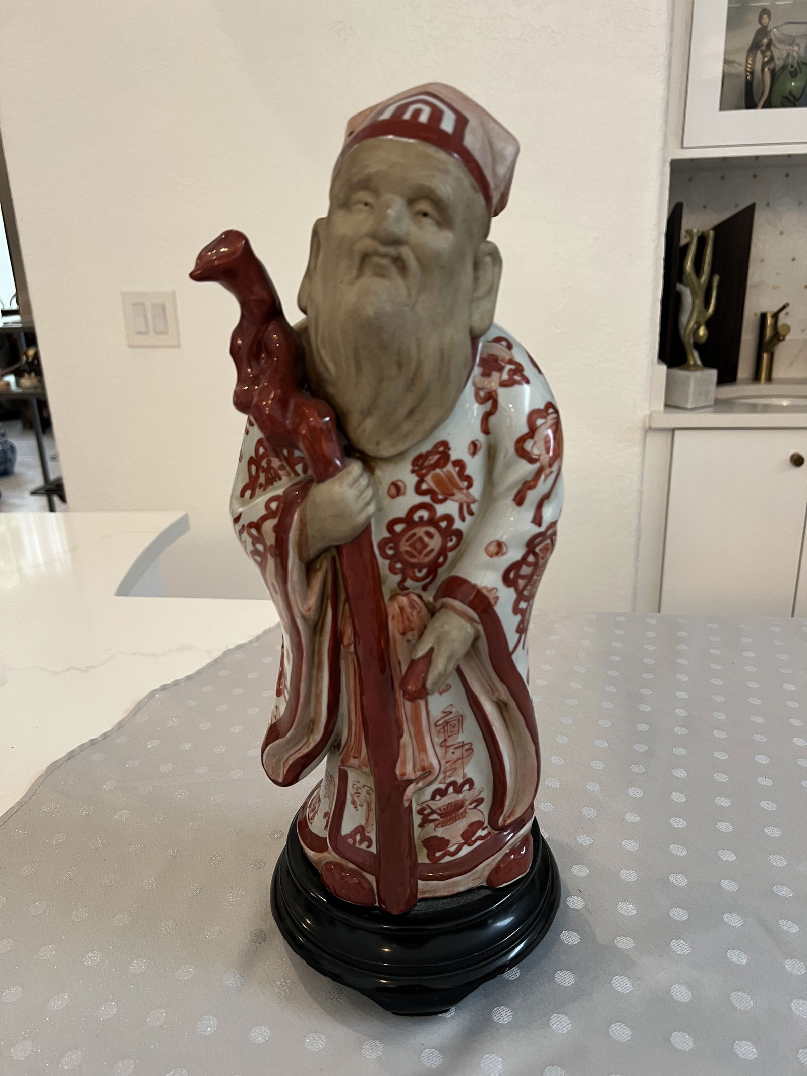 Japanese God Statue: Porcelain figure on wooden base, rust or cinnabar color, with walking stick or cane. Highly decorative. Dimensions: H 17" x W 6" x Dia: 6" Condition: Presenting well. Low Estimate: 100.00High