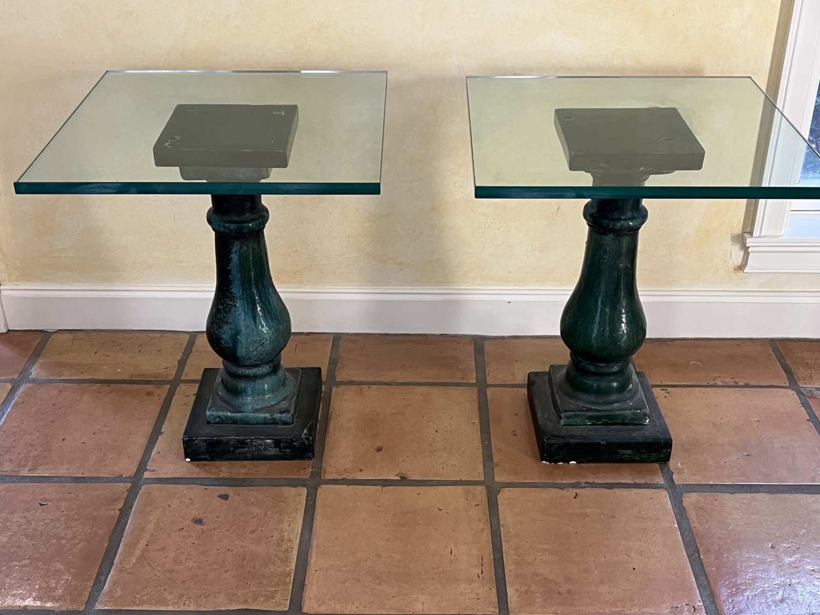 Pair of Green Glazed Pillar Tables: Blue green glazed pottery pedestal tables, glass tops. Dimensions: H 23.5" x W 9" (base) x 20" (top) Glass: almost 1" Condition: Repair to one corner, large chip to another, smaller losse to bottom