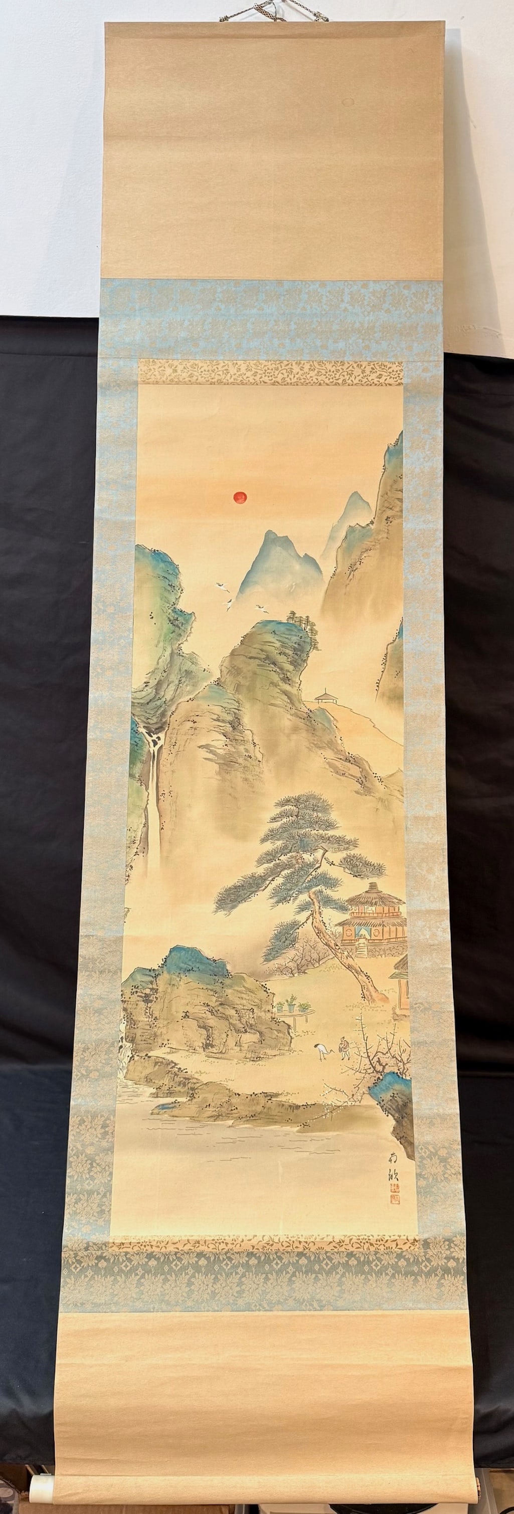 CHINESE LANDSCAPE HANGING SCROLL, SIGNED FANG XIN, 20 th CENTURY, 20IN x 75IN (51cm x 191cm): A finely rendered Chinese landscape painting in the classical shan shui (mountain-and-water) tradition, depicting a tranquil mountain retreat scene beneath a rising red sun. The composition features l