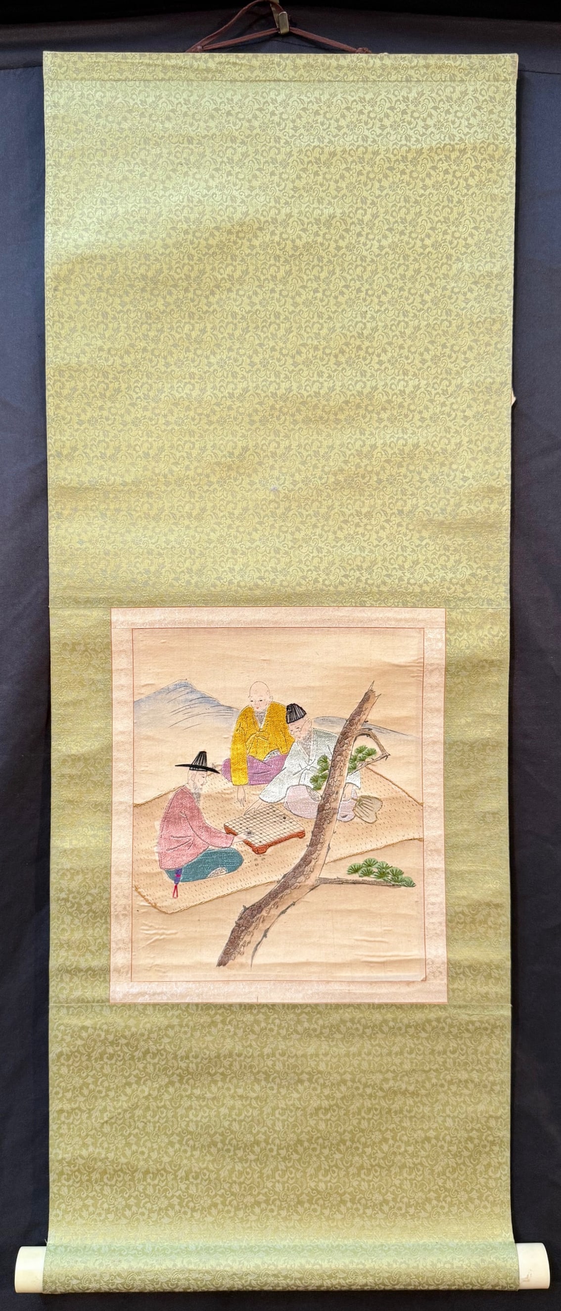 CHINESE EMBROIDERED SCROLL OF THREE MEN PLAYING WEIQI, 12.5IN x 33IN (32cm x 84cm): A Chinese embroidered scroll of three men playing Weiqi. It is adhered to a silk backing. The embroidered silk square measures 8 inches x 11 inches. The entire scroll measures 12.5 inches X338 inches.