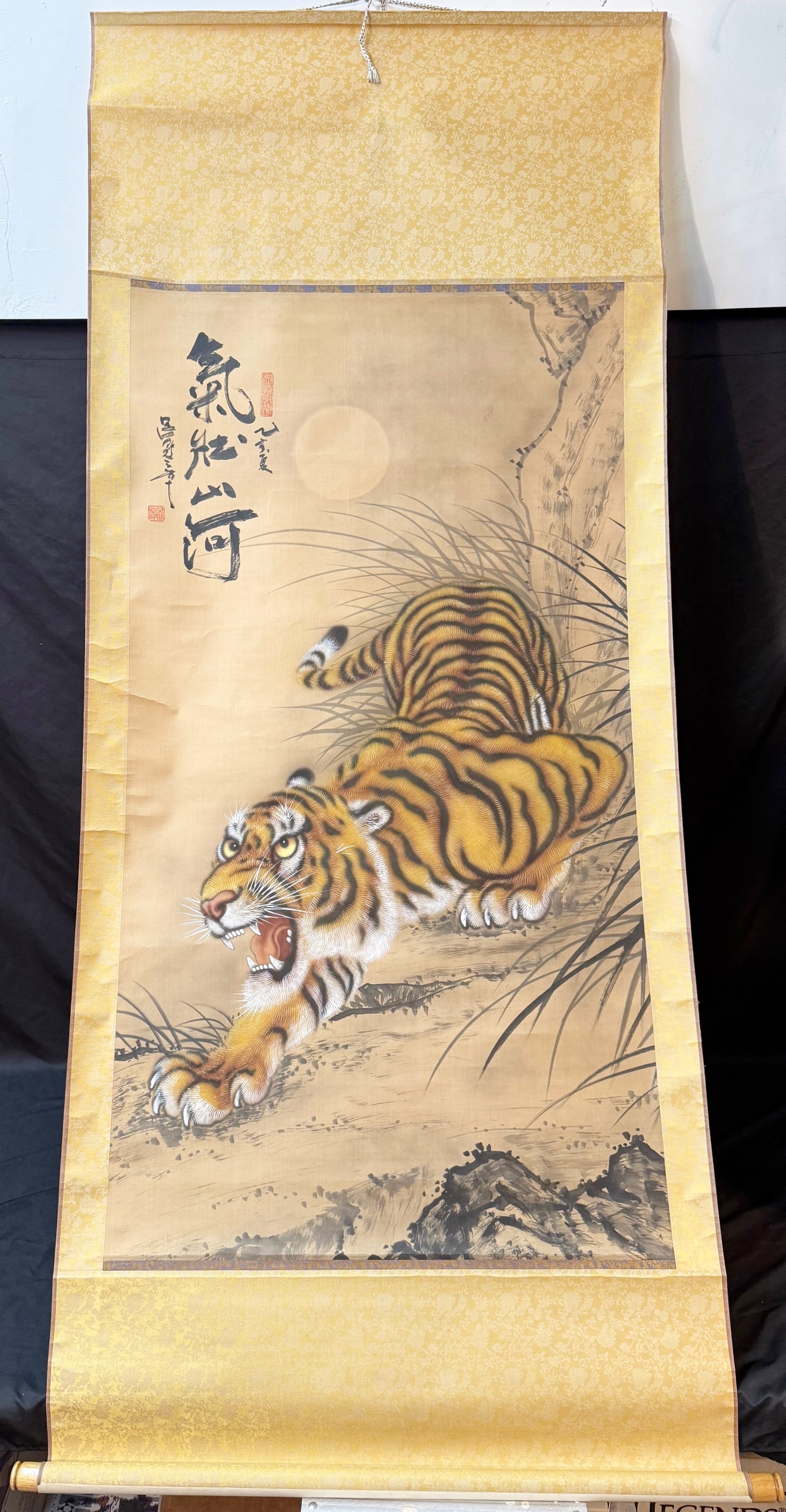 ANTIQUE JAPANESE INK AND COLOR HANGING SCROLL OF A TIGER, 32IN x 72IN (81cm x 183cm): A finely executed Japanese hanging scroll depicting a?crouching tiger emerging from tall grasses beneath a luminous full moon, rendered in expressive ink and mineral pigments on silk. This scroll was 