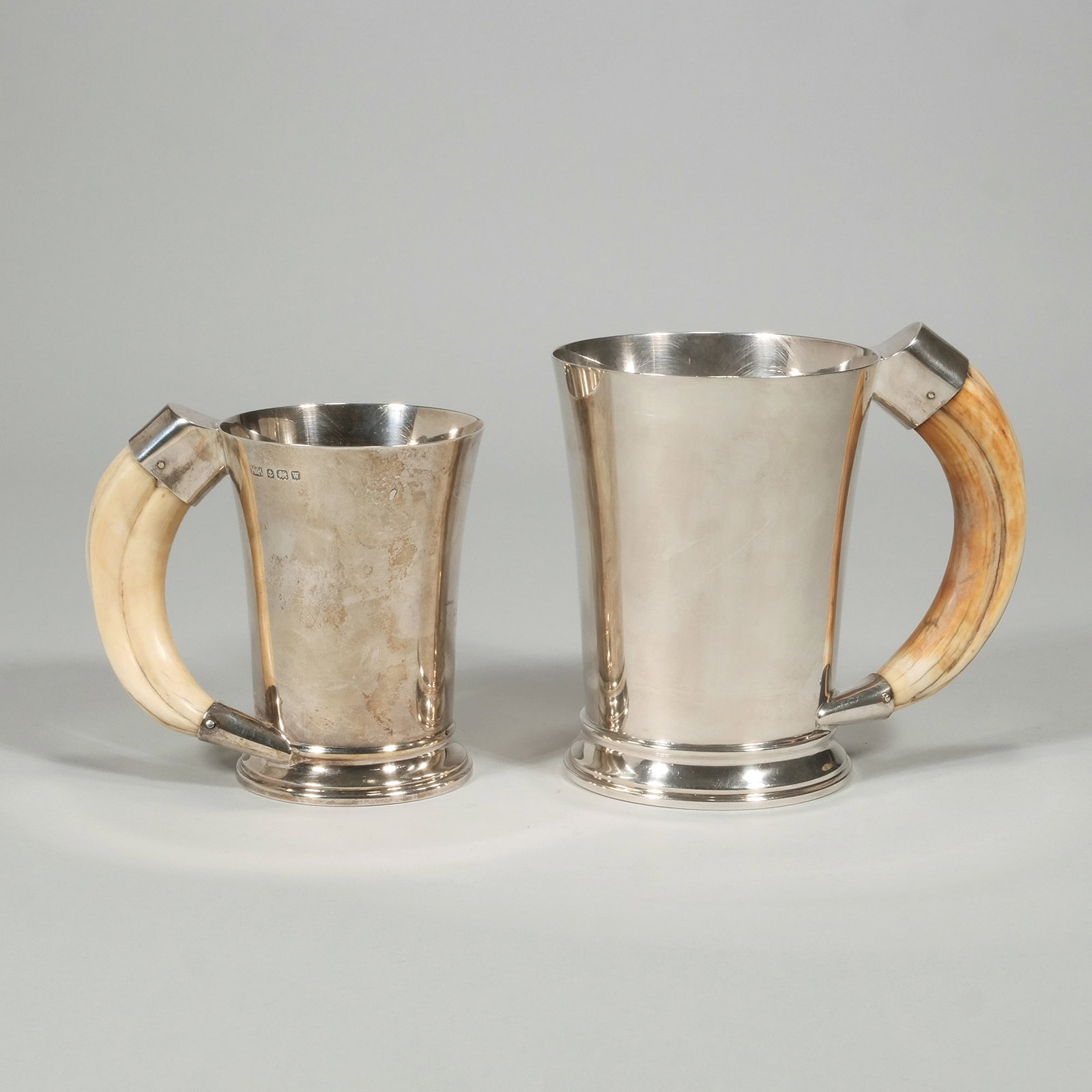 (2pc) ANTIQUE ENGLISH STERLING SILVER BOAR HANDLE HUNT MUGS "HH": Hunting mugs with boar tusk handles, the smaller mug having a 5 Shilling piece set into the bottom total weight 29.4 oxt h. 5.5 x dia. 4 in. (Taller) Provenance: Items from the private collection of m