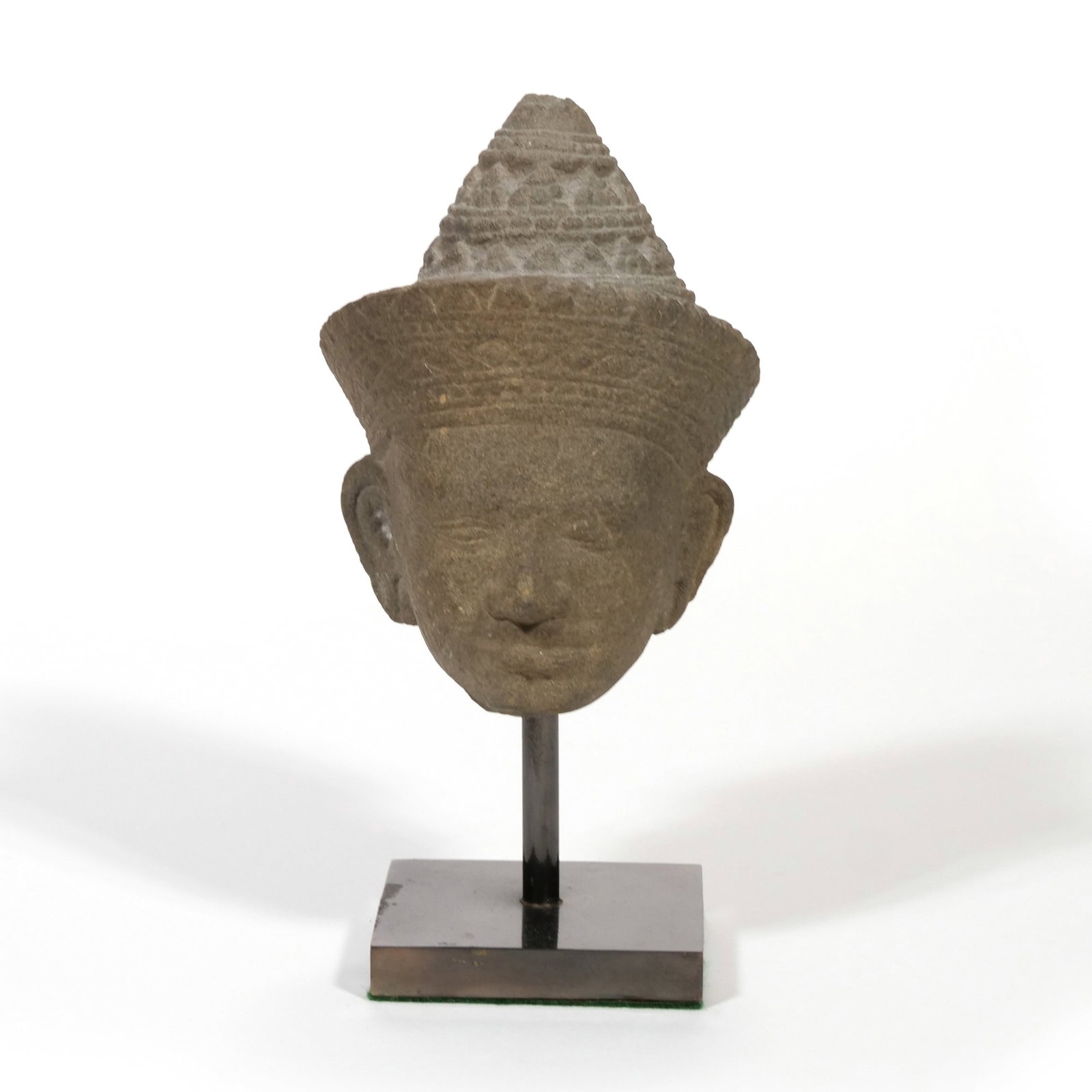 KHMER KOH KER SCHOOL SANDSTONE SCULPTURE: Head of a Deity Carved sandstone h. 7.5 x w. 3.5 in. Circa 10th century, the head features a tiered conical headdress with geometric patterns and elongated earlobes, mounted on a square black metal st
