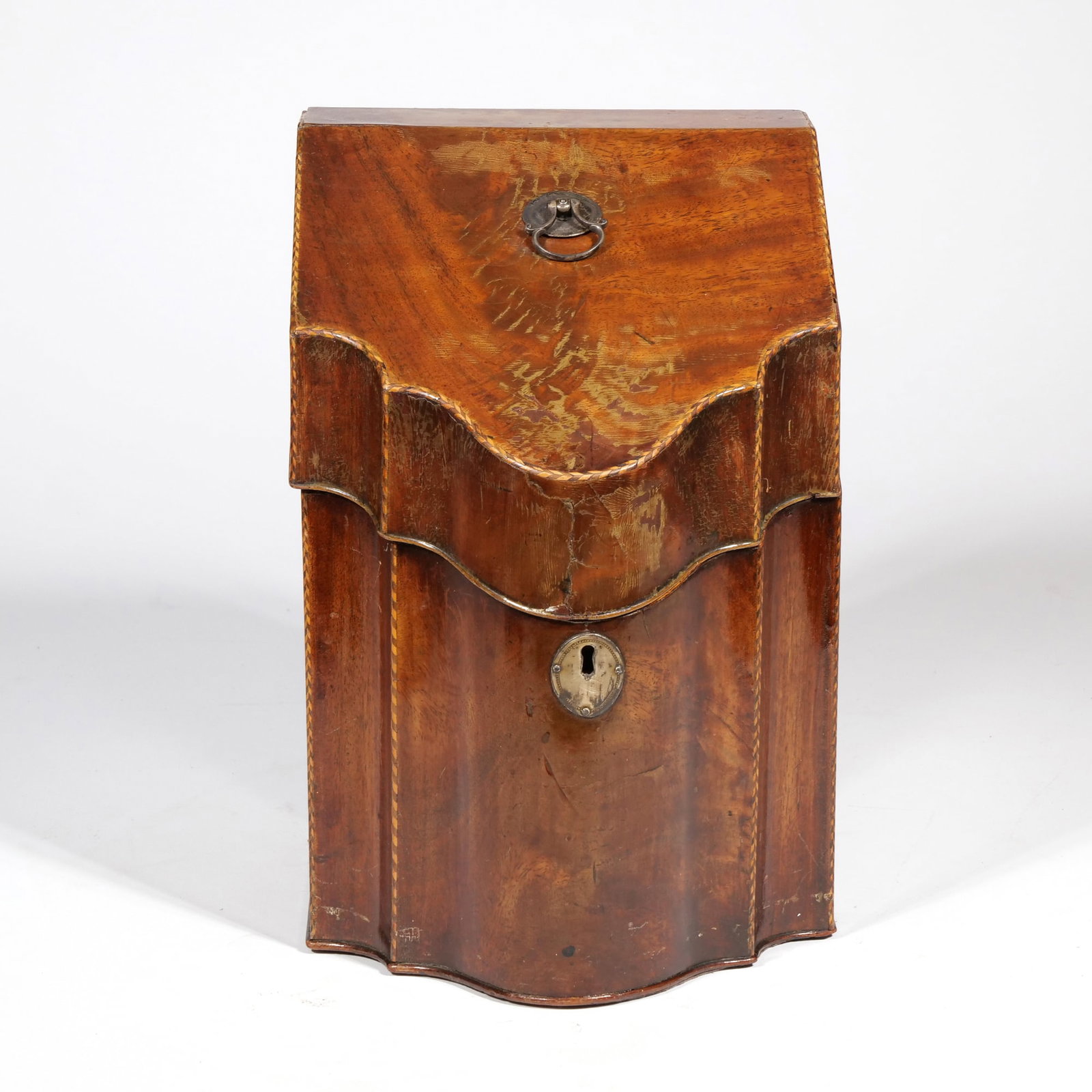 GEORGE III MAHOGANY SERPENTINE KNIFE BOX: Constructed from mahogany with a sloped lid and a serpentine-front profile. The exterior features checkered stringing along the edges and a silver metal escutcheon and lid ring pull (with 'F' hallmark