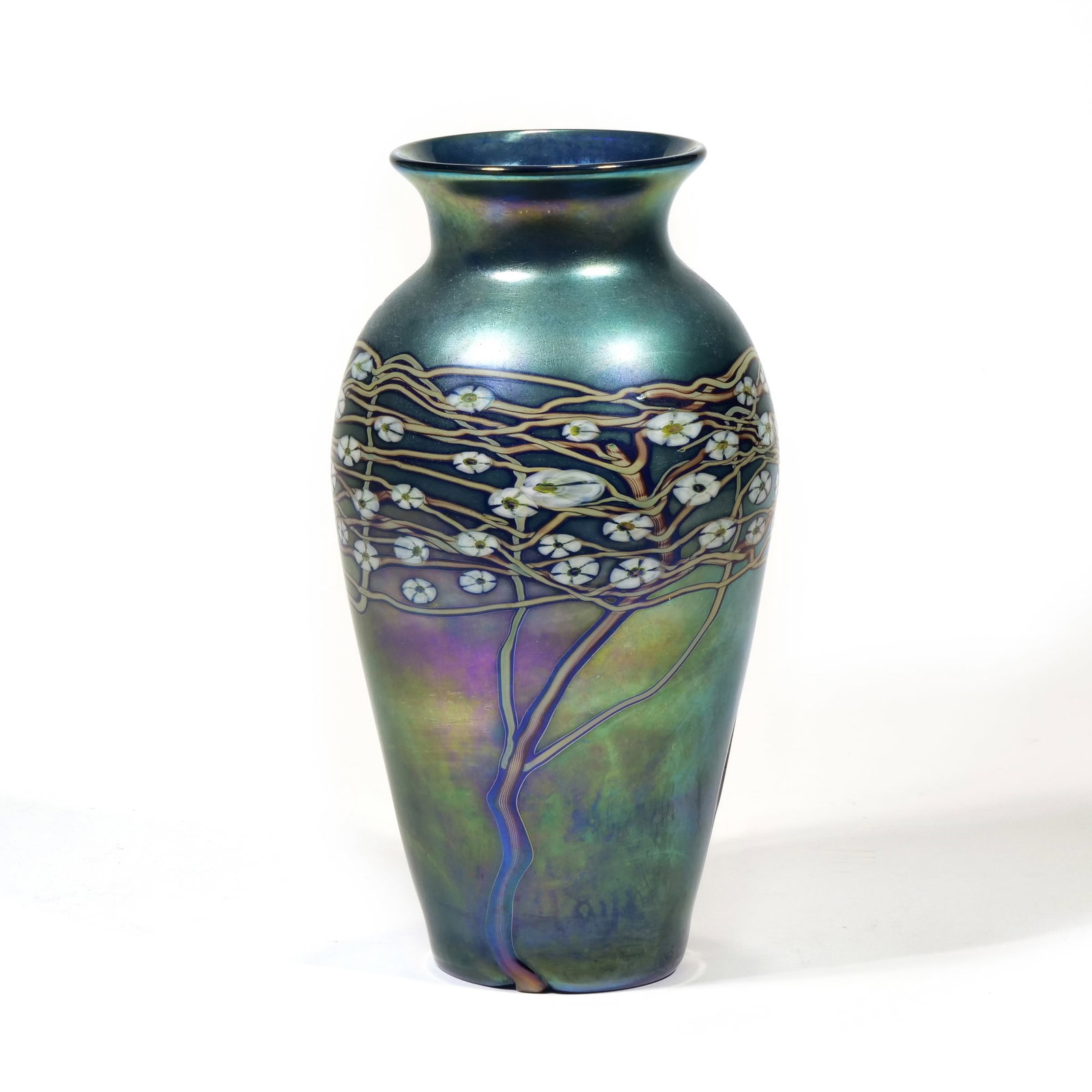 ZELLIQUE STUDIO IRIDESCENT ART GLASS VASE: The iridescent art glass vase features a tapered form with a flared rim and a teal to green gradient background. A decorative band of white blossoms on intertwined brown branches encircles the upper b